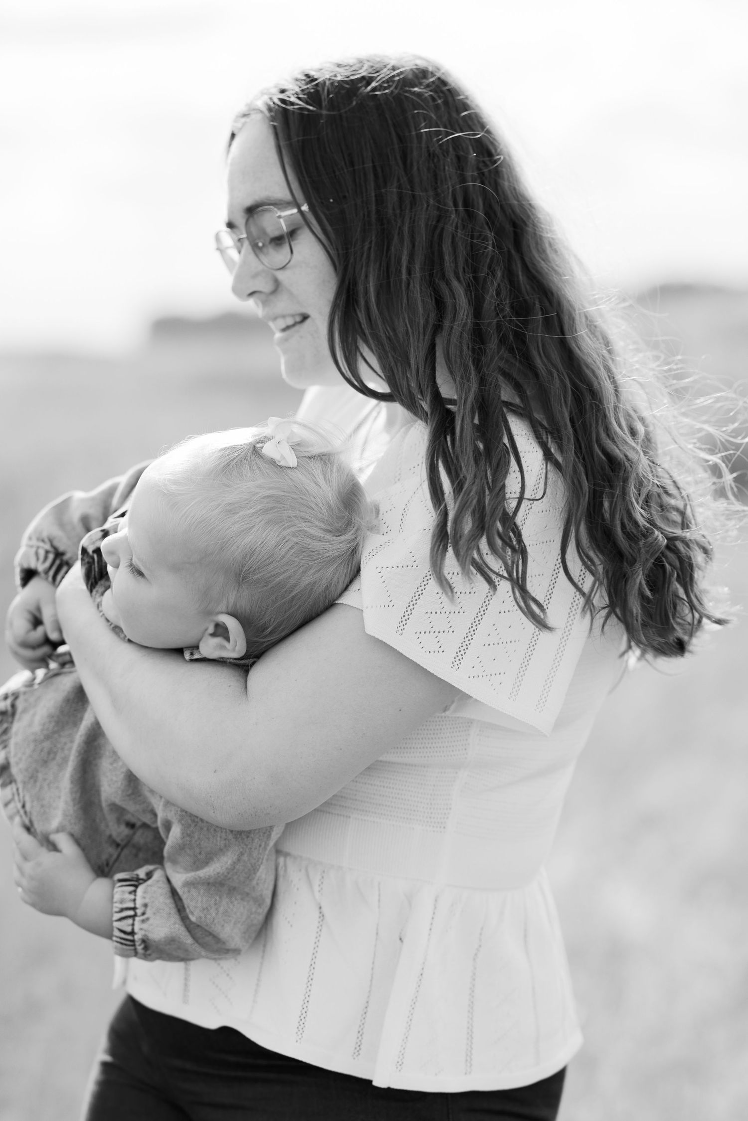 Black and white photo of a smiling woman with glasses holding a sleeping baby outdoors in a field.