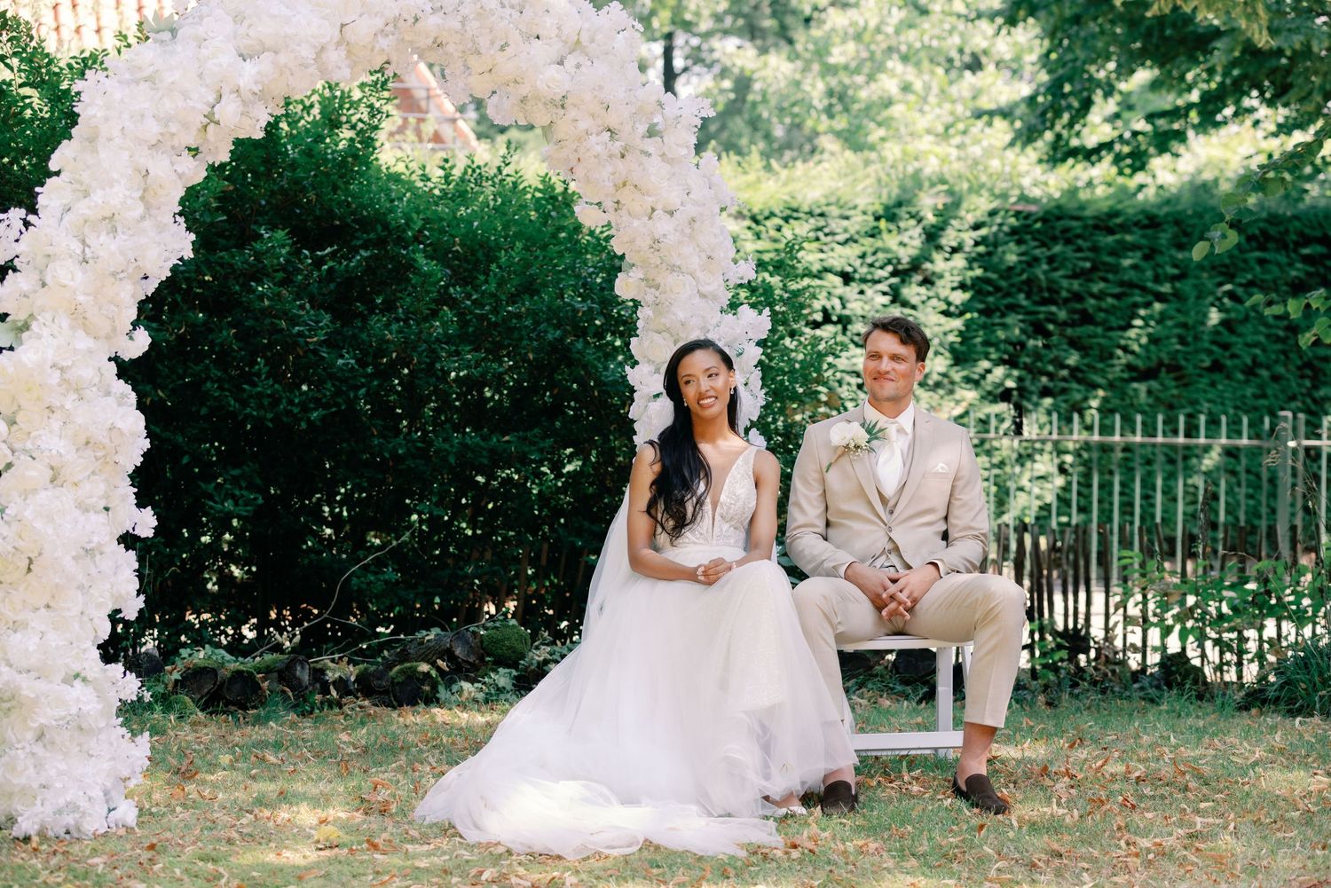 A couple in wedding attire poses together under a decorative white floral arch in an outdoor garden setting.