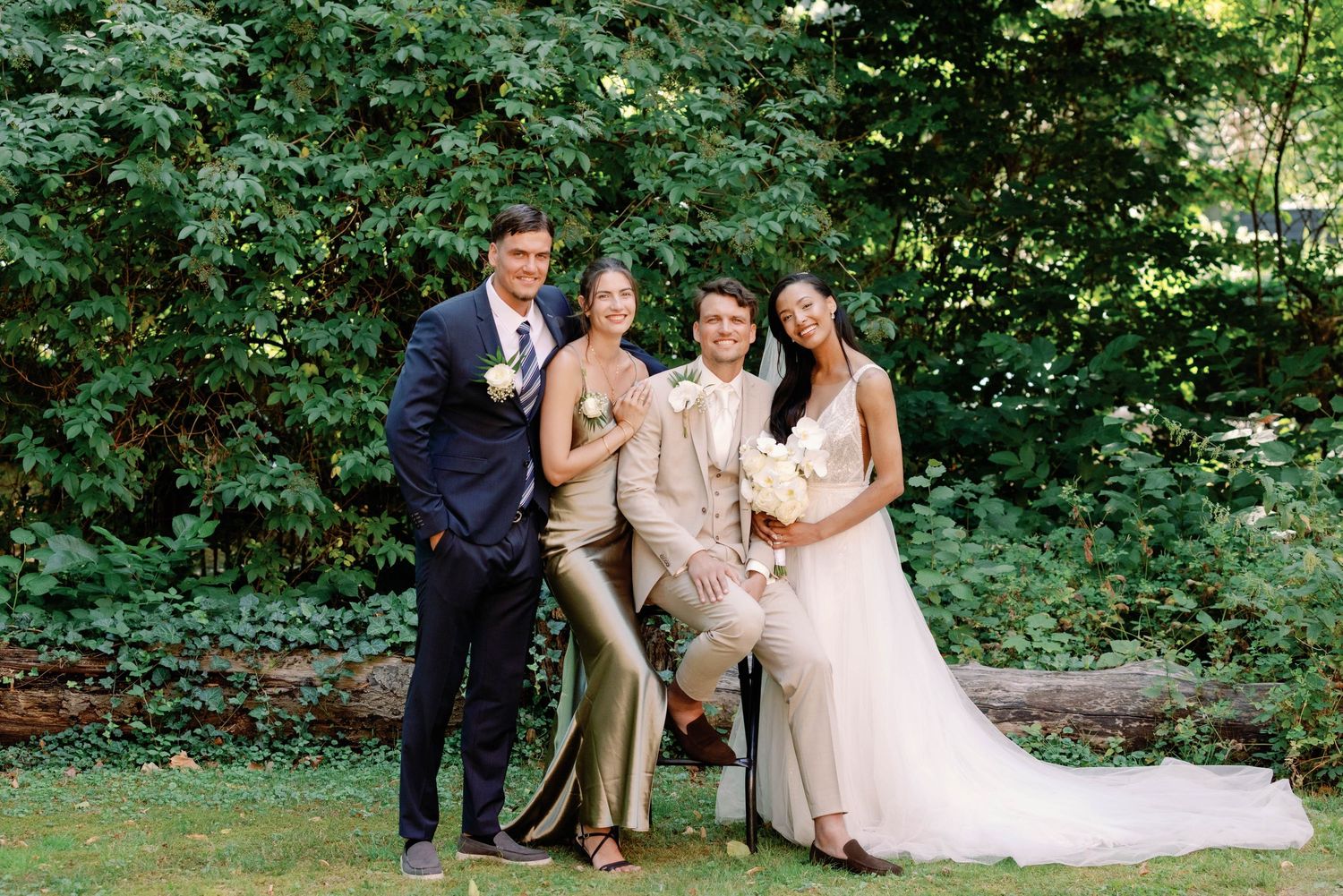 A wedding party poses together in a lush green forest setting with sunlight filtering through the trees.
