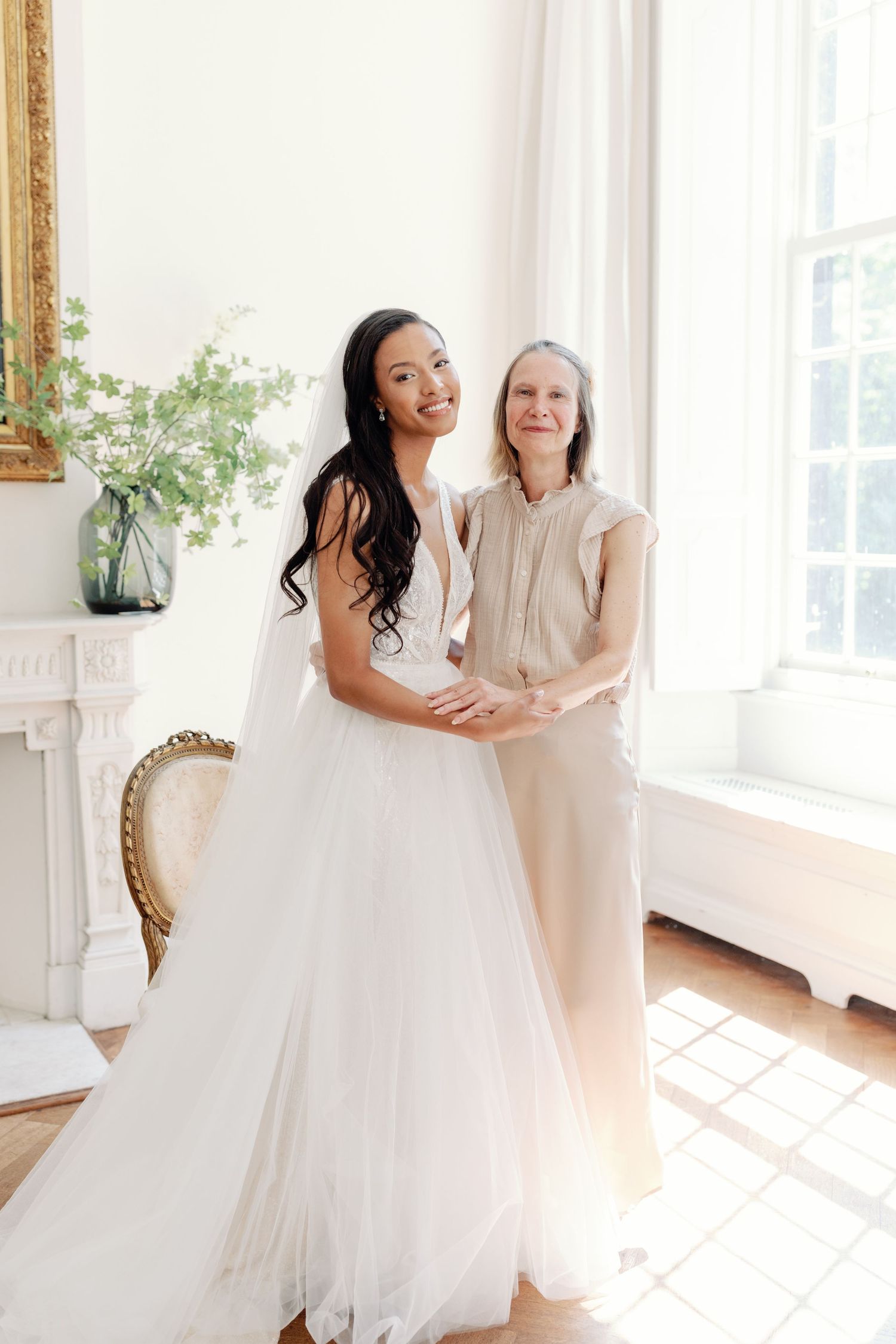 Two people share a tender moment in a bright sunlit room with a white wedding dress and elegant decor.