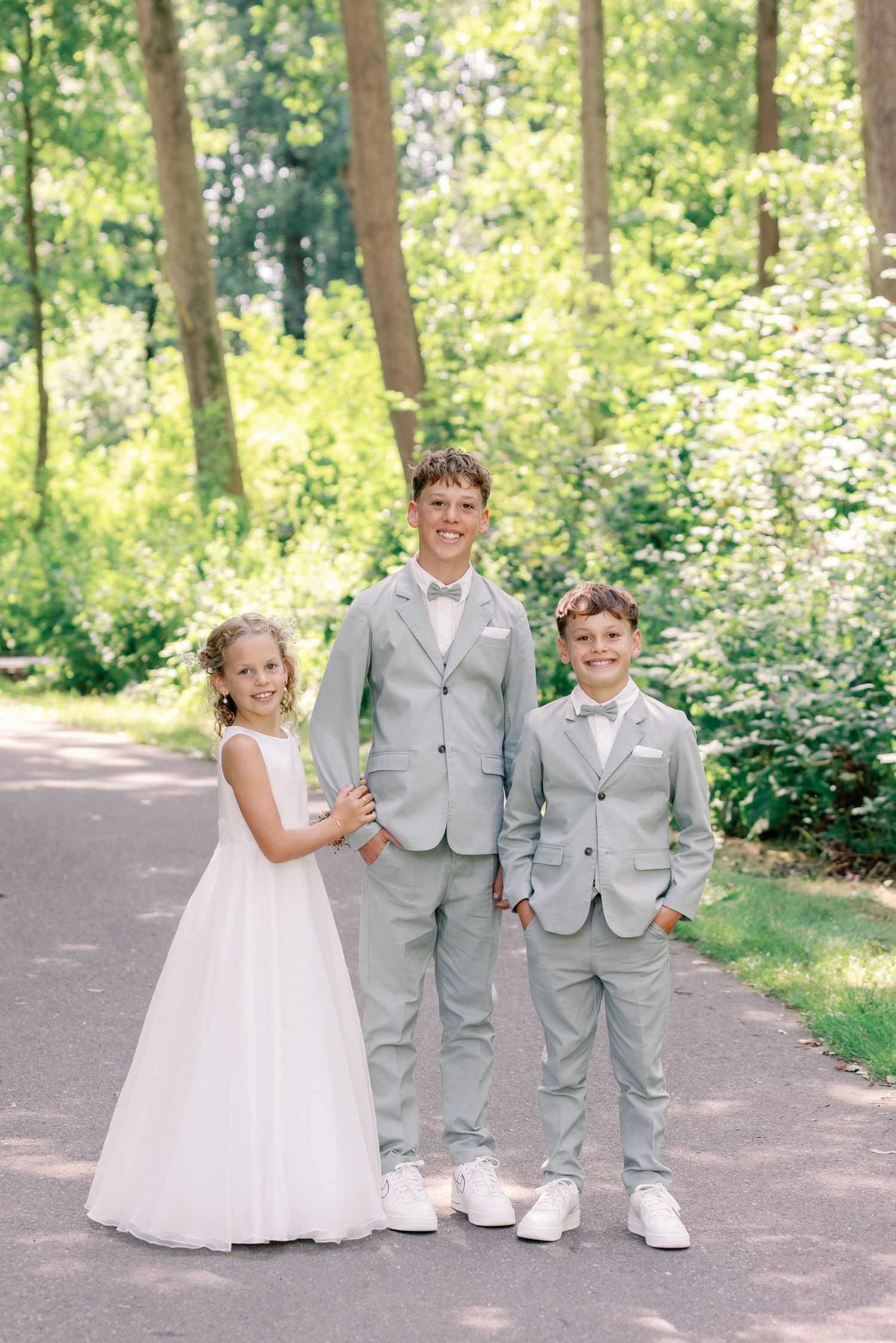 Children in formal wedding attire pose playfully on a wooded path during a summer celebration.