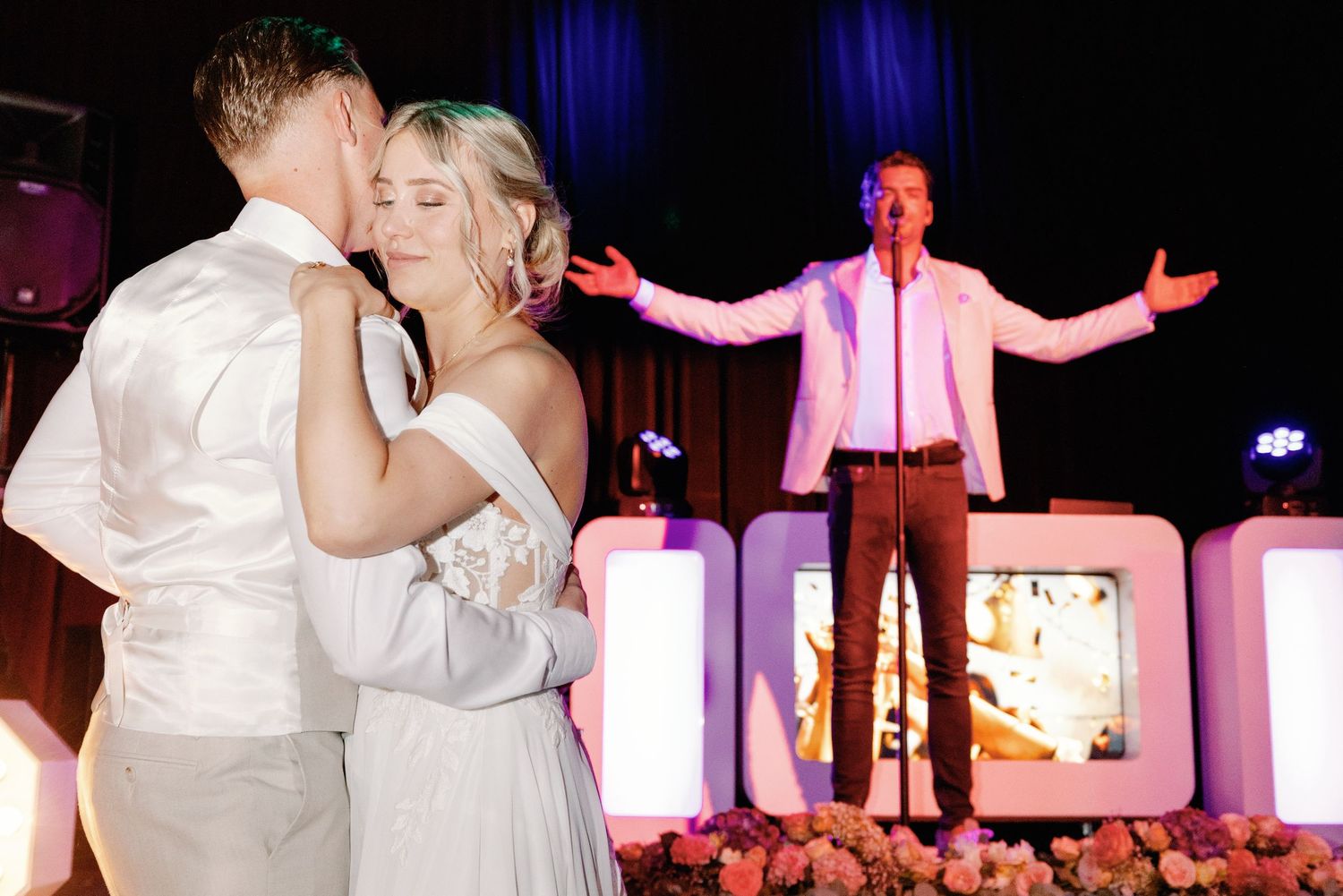 A couple shares a romantic dance at their wedding reception while an entertainer performs on stage in the background.