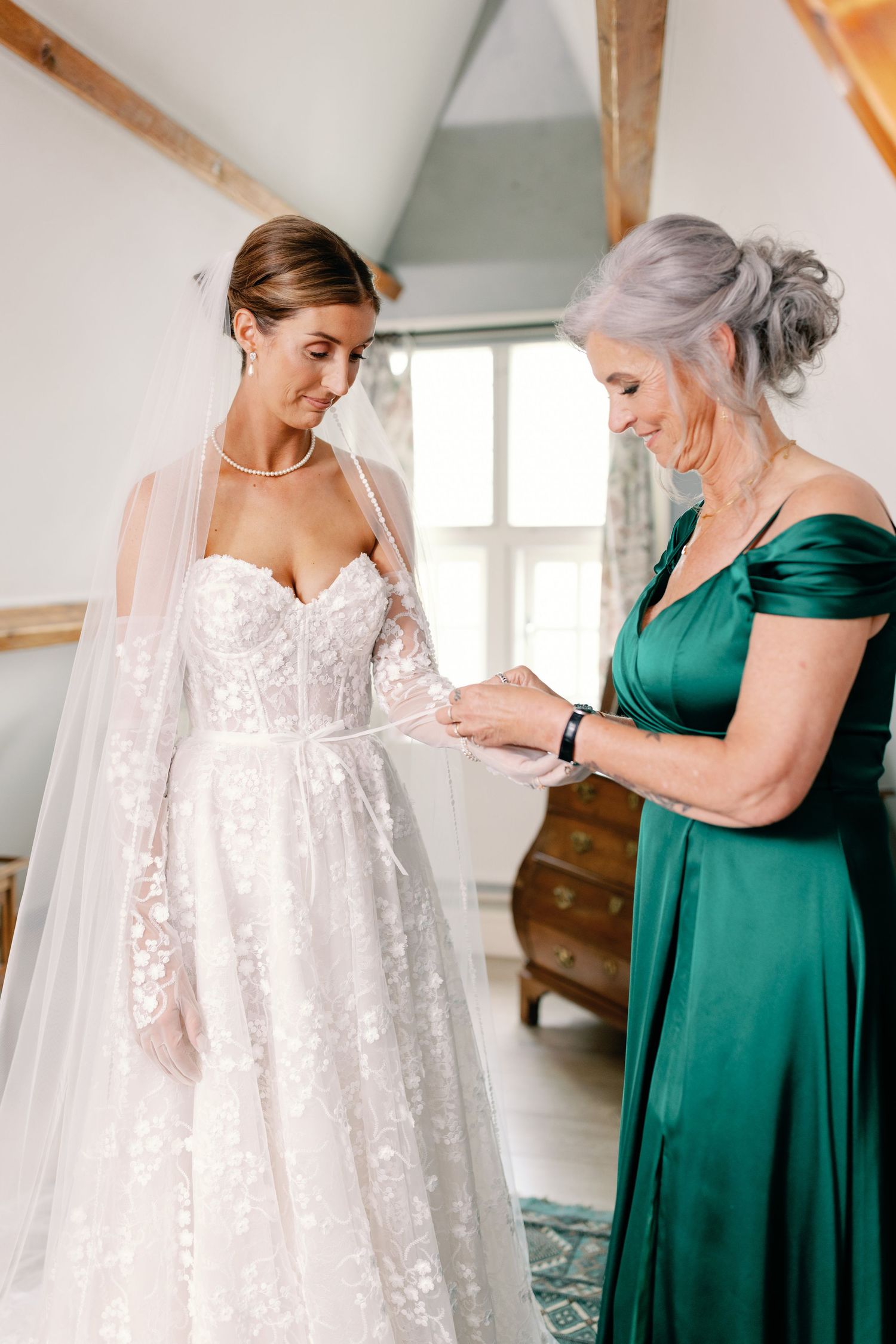 Two women share a special moment as they prepare for a wedding, with one wearing a lace gown and the other in emerald green.