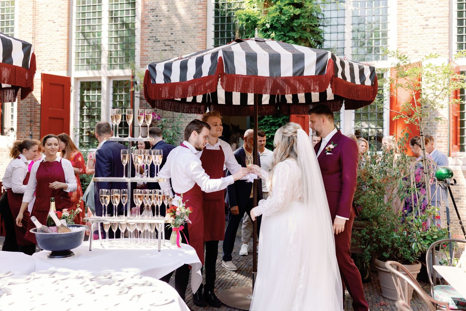 An outdoor wedding ceremony under a striped awning with burgundy and white decor in a garden setting.
