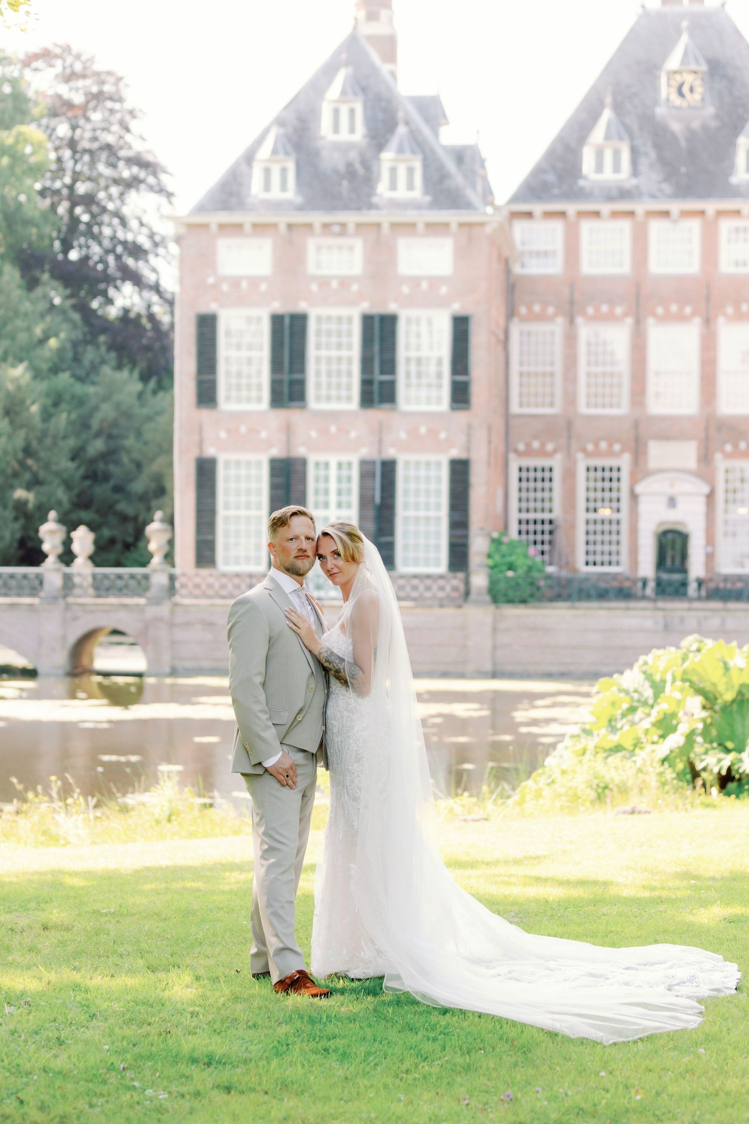 A romantic wedding couple poses together on a manicured lawn in front of a grand historic brick mansion.