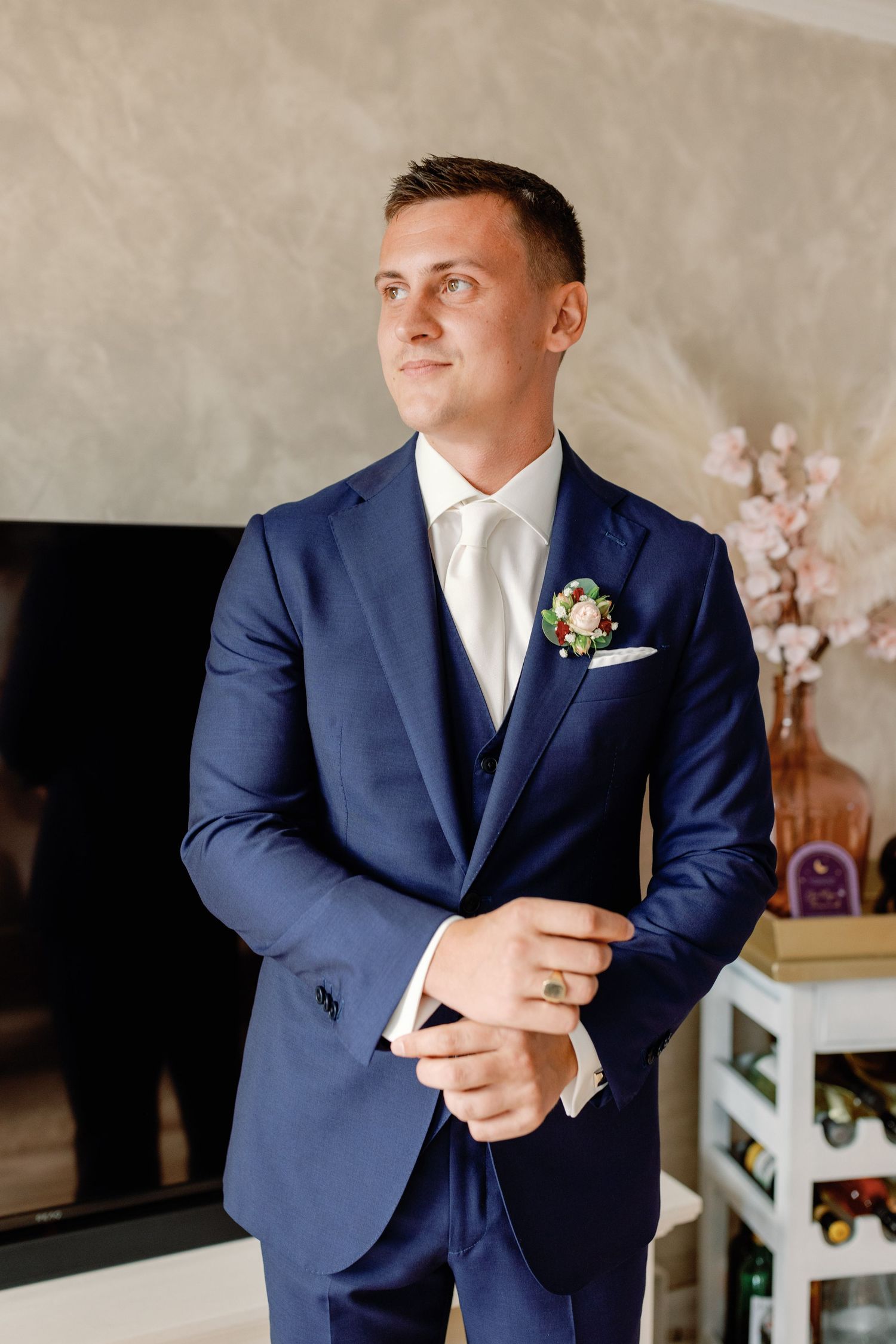 Groom in classic navy suit poses against neutral wall during wedding preparations.