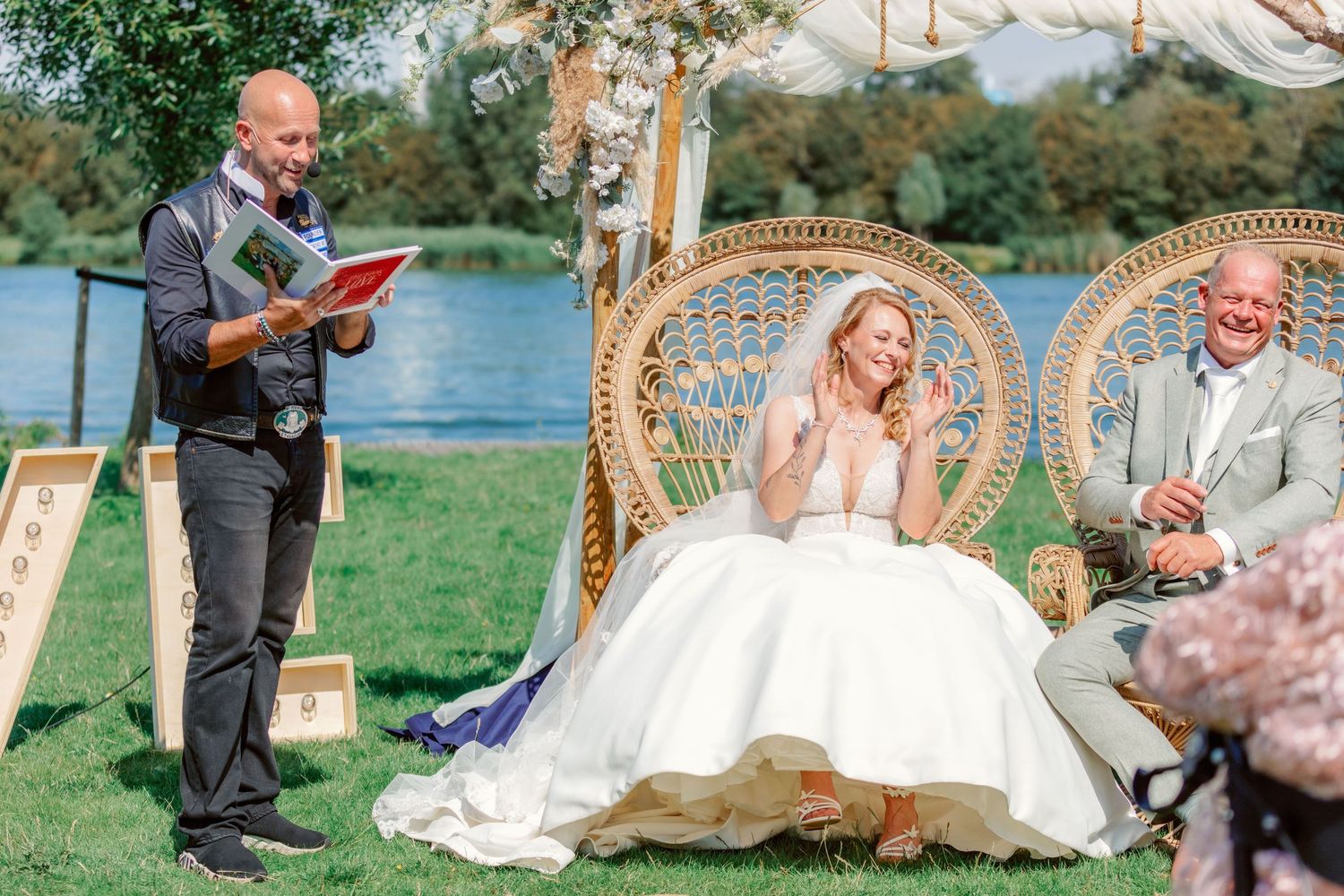 Outdoor wedding ceremony by a lake with peacock chairs and floral arch decorations.
