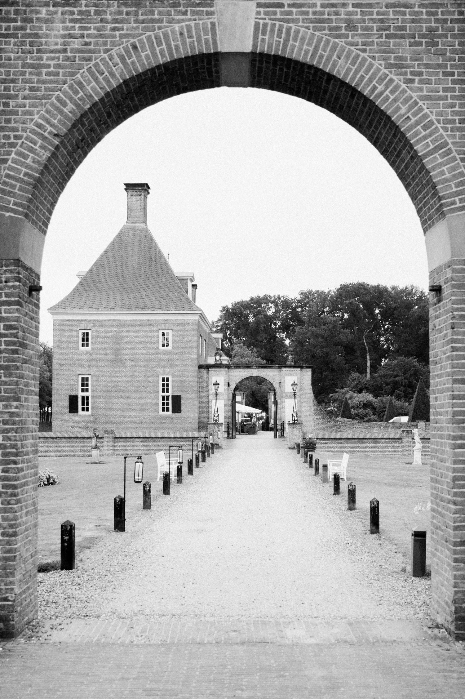 Black and white view through brick archway showing historic building entrance with bollards lining gravel path.