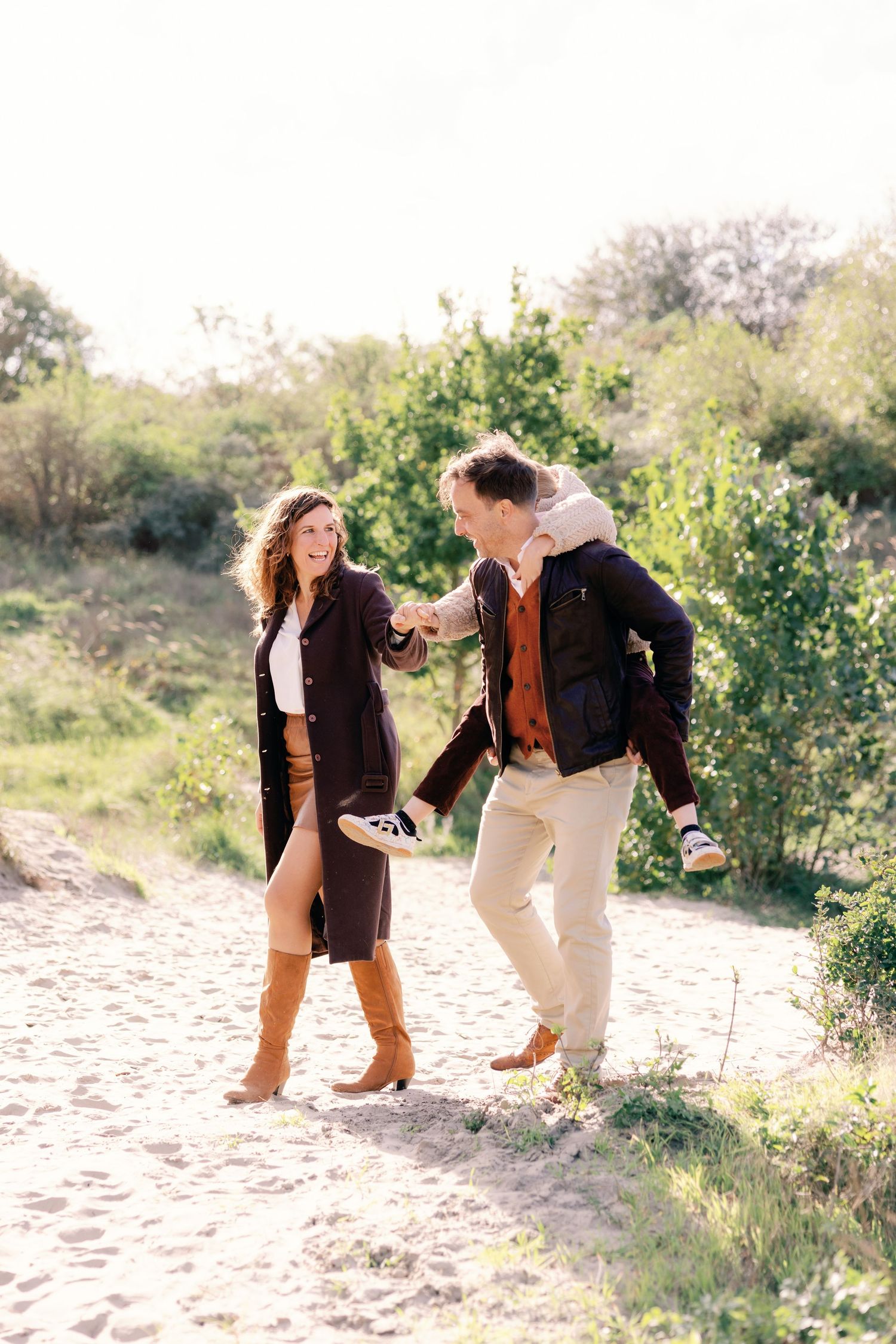 Couple holding hands and dancing on a sunny sandy path surrounded by green bushes outdoors.