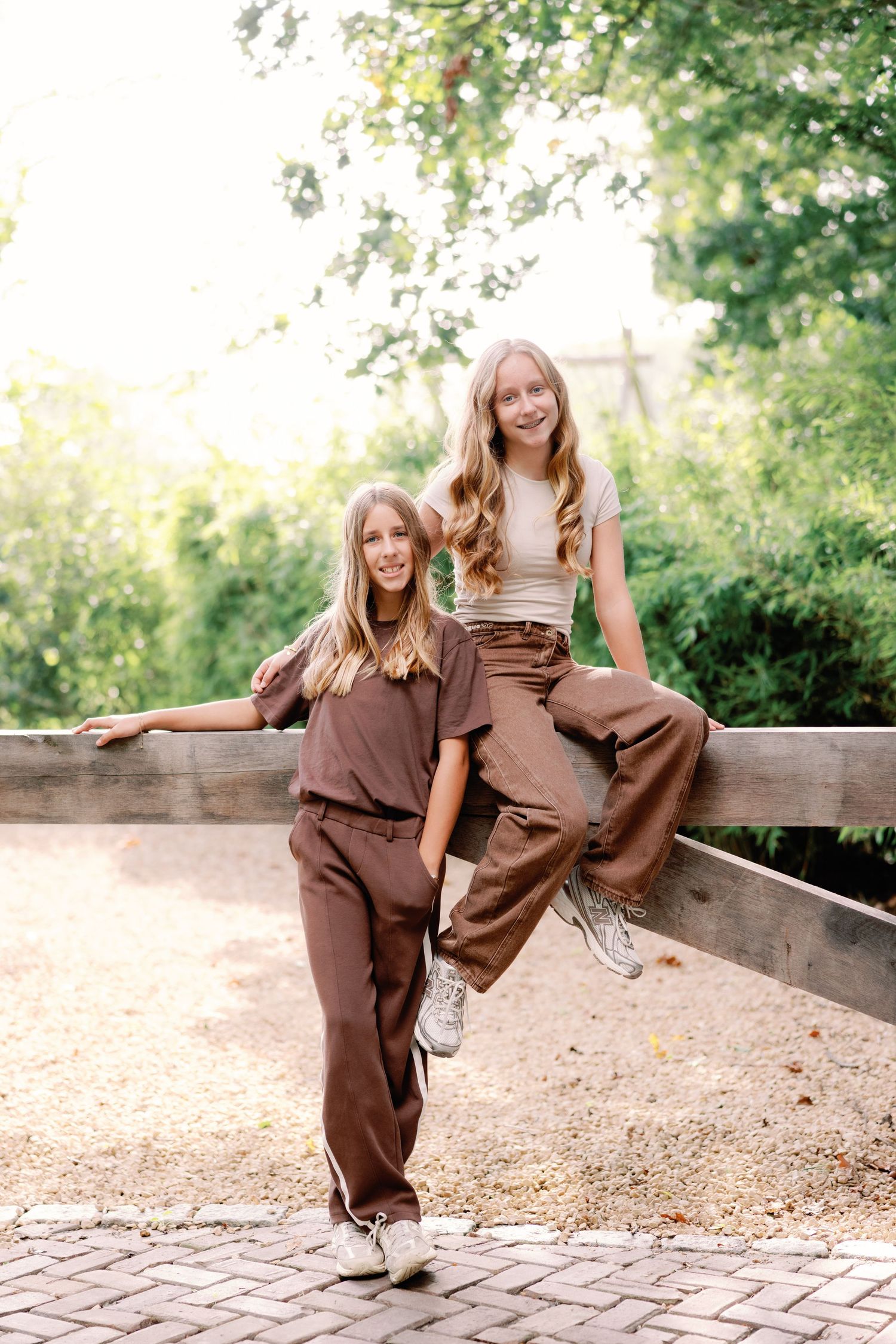 Two girls in matching brown outfits and sneakers pose on a wooden fence beside a brick path, framed by sunlit green trees.