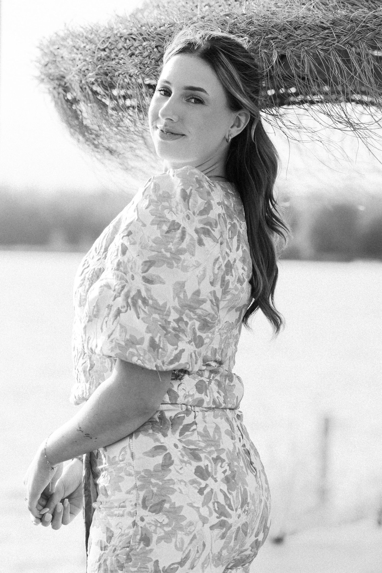 Black and white photo of a smiling woman in a floral dress posing outdoors under a straw umbrella.