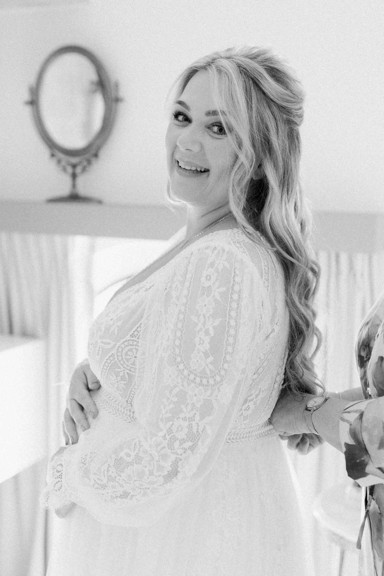 Black and white portrait of a bride in a lace wedding dress smiling over her shoulder with a vintage mirror in background.