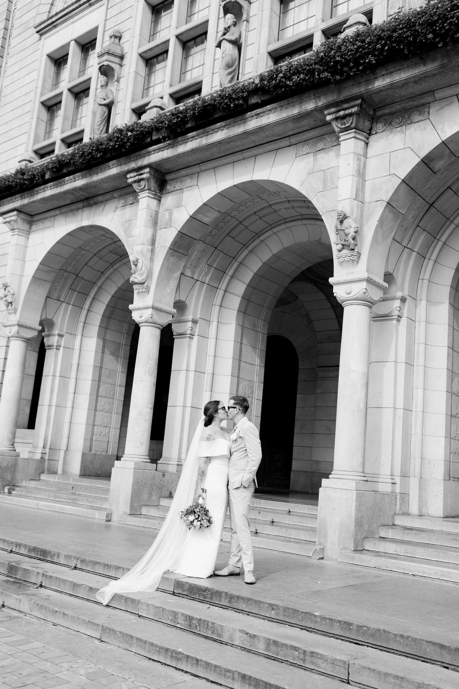 A romantic couple shares a tender moment under the arched columns of a grand historic building.