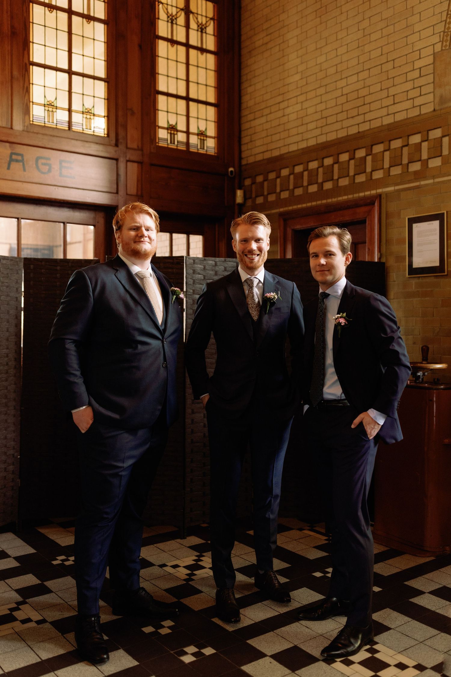 Group of businessmen in dark suits standing in a hallway with checkered floor and brick walls.