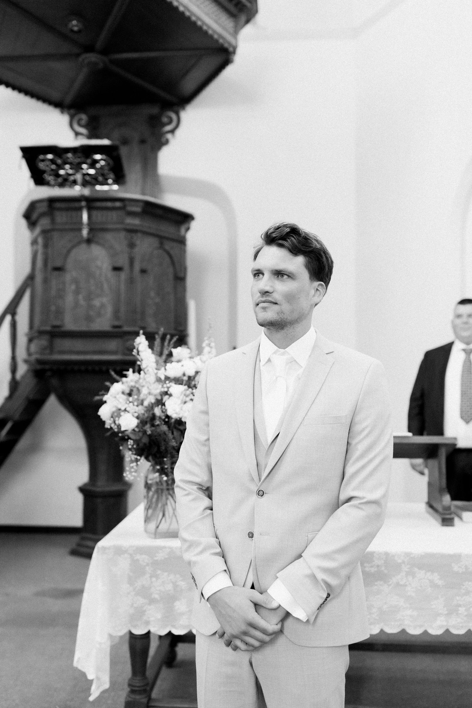 A groom in a light colored suit stands thoughtfully during an elegant outdoor wedding ceremony.