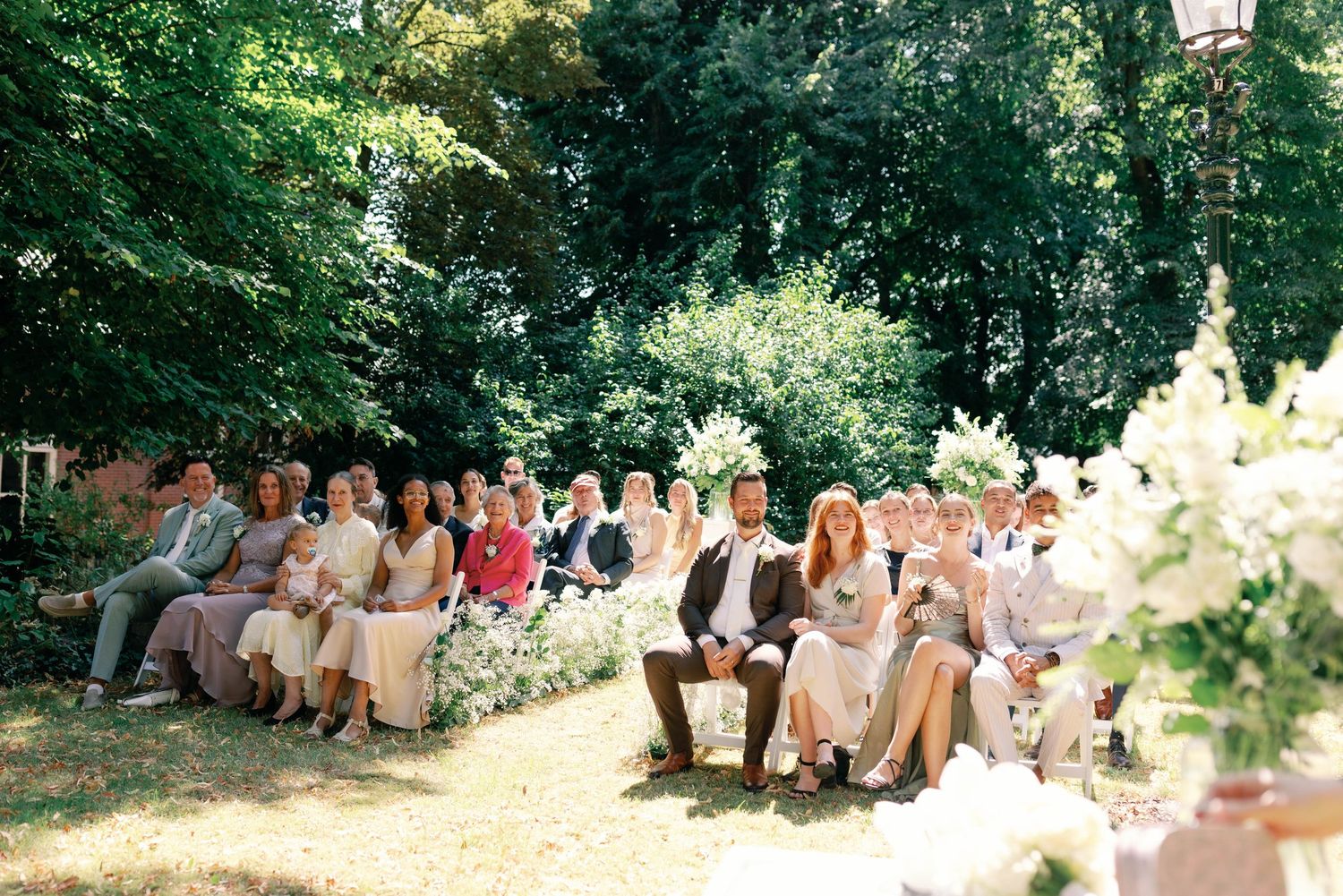 Wedding guests seated outdoors in a garden setting with trees and sunlight filtering through the foliage.