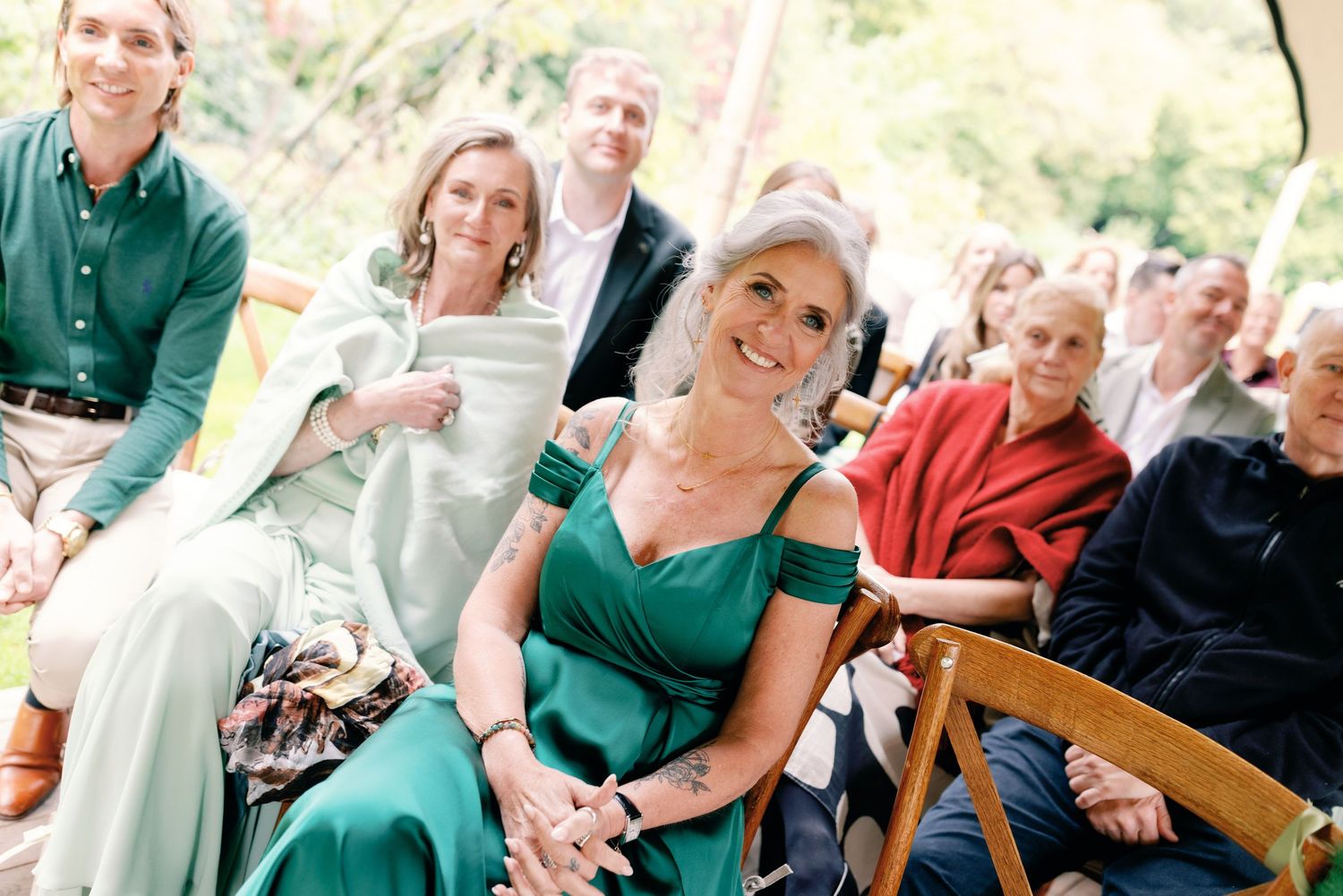 Wedding guests seated in wooden chairs at an outdoor ceremony with someone wearing an elegant emerald green dress.