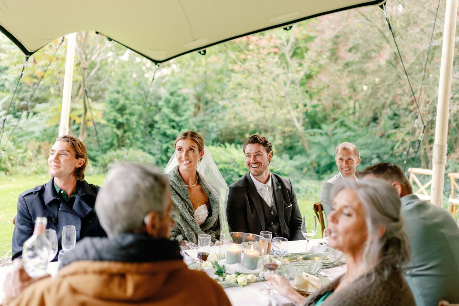 A group of people enjoying a meal together at an outdoor covered dining area surrounded by lush greenery.