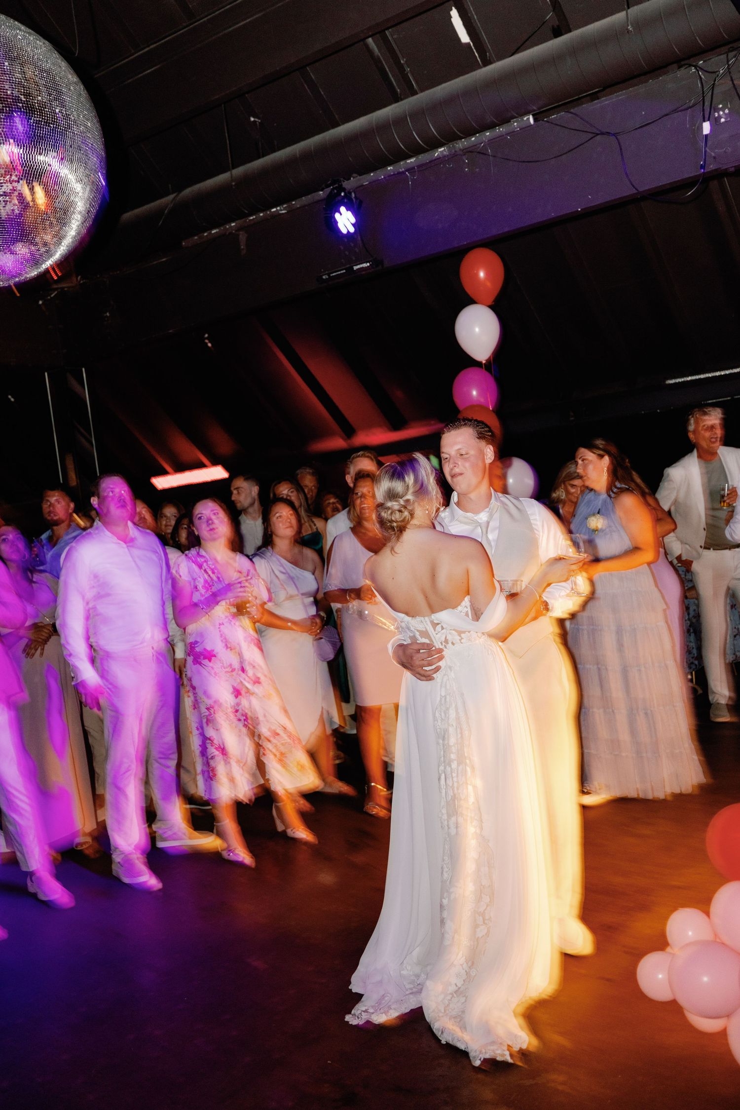 Wedding guests dance together in white attire under purple lighting at an indoor reception venue.