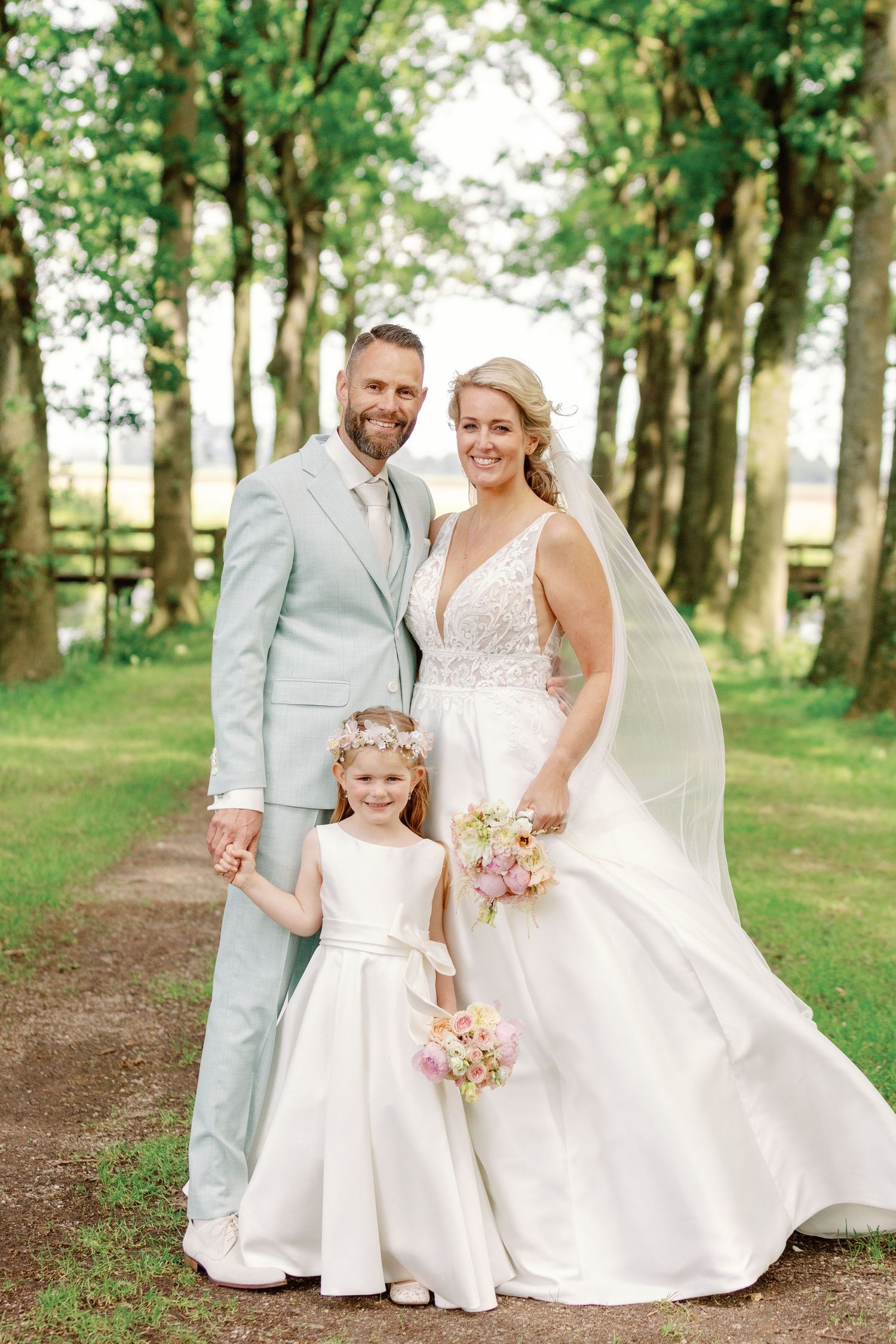 A wedding couple and young flower girl pose together on a tree-lined path in elegant white attire.