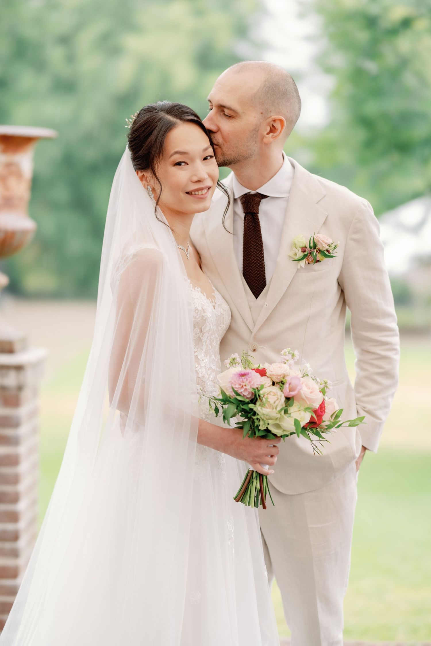 A couple in wedding attire share an intimate moment outdoors with the bride holding a pink and white bouquet.