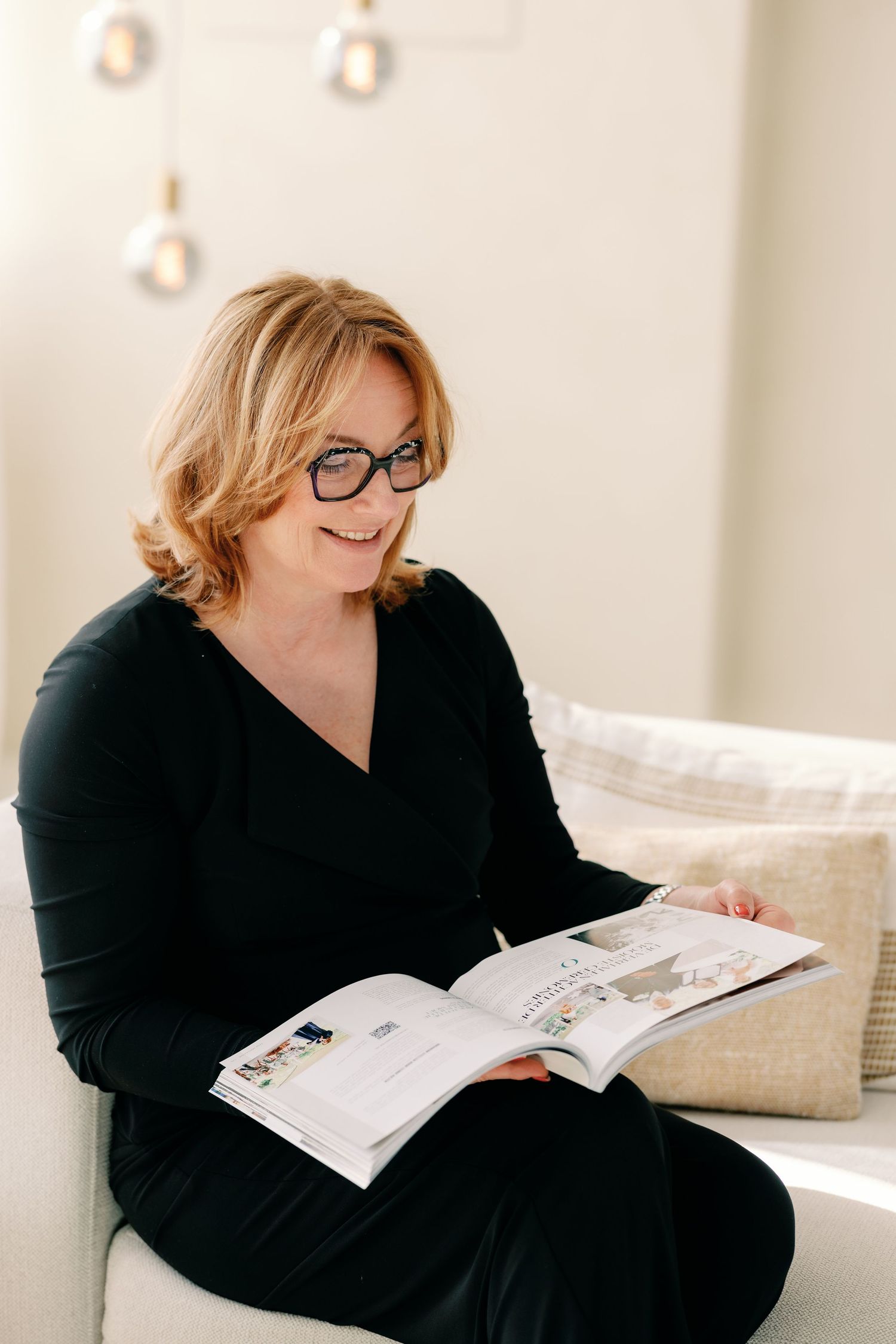 A person wearing black lounges on a couch while reading a magazine in a bright, minimalist interior setting.