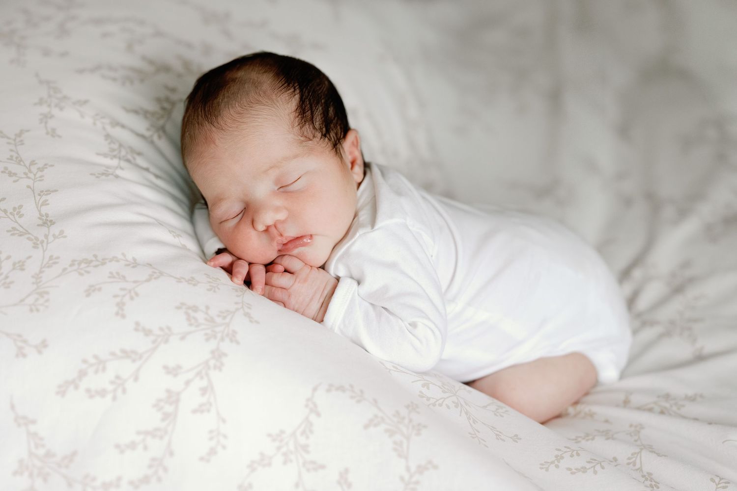 A sleeping newborn baby dressed in white rests peacefully on soft white bedding.