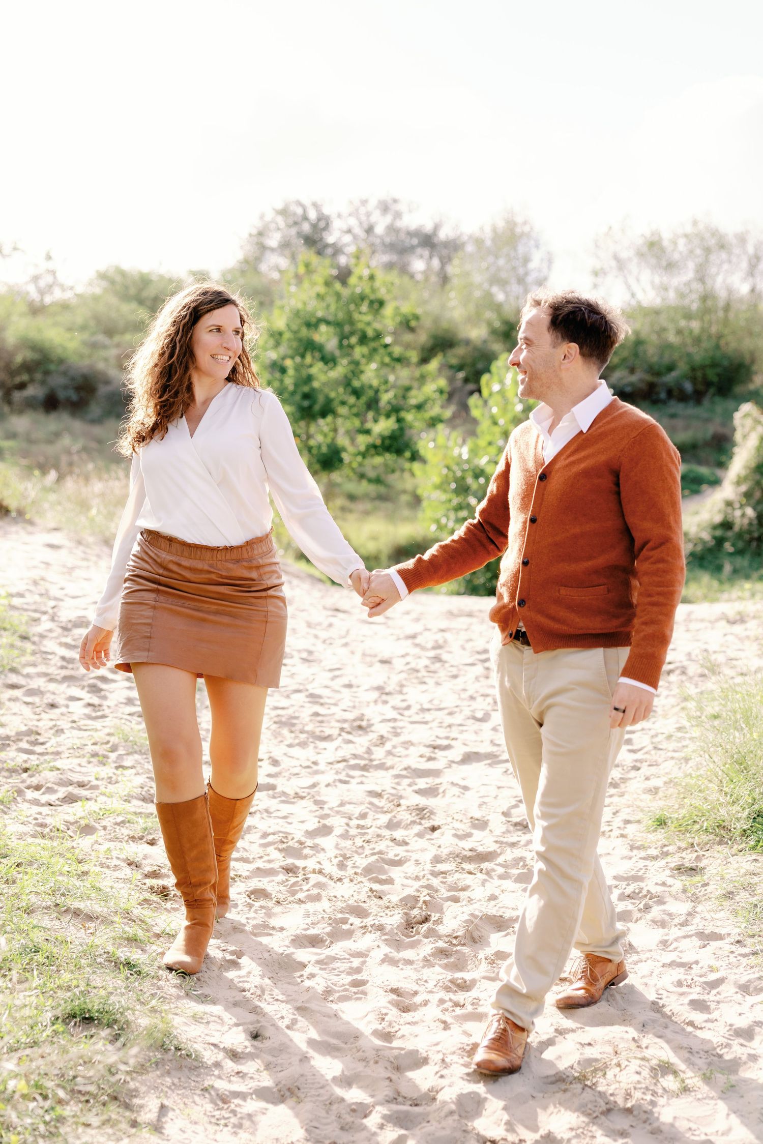 Couple holding hands and walking on a sandy path surrounded by greenery in warm autumn outfits.