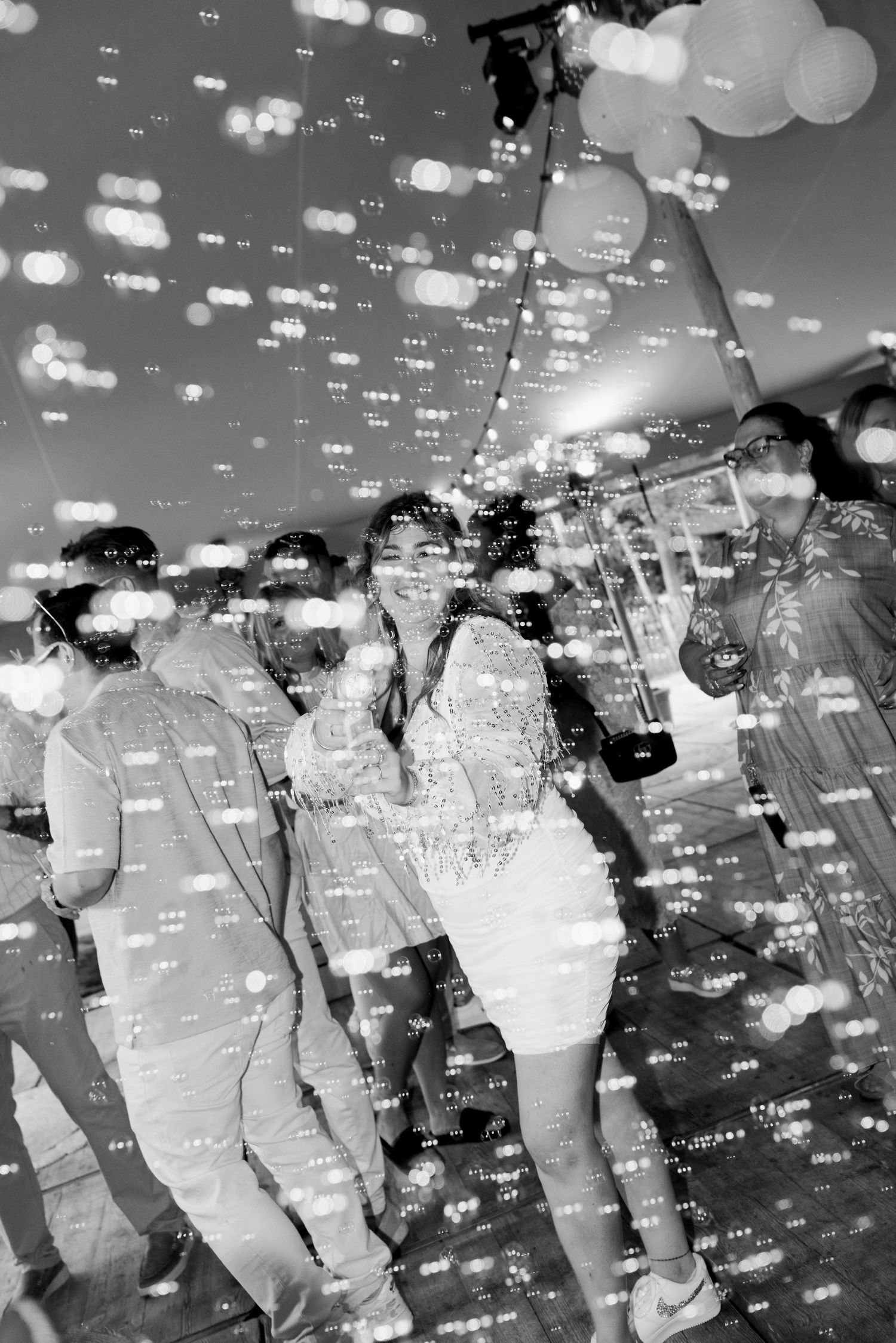 Black and white photo of smiling woman dancing at a festive party with bokeh lights and balloons in background.