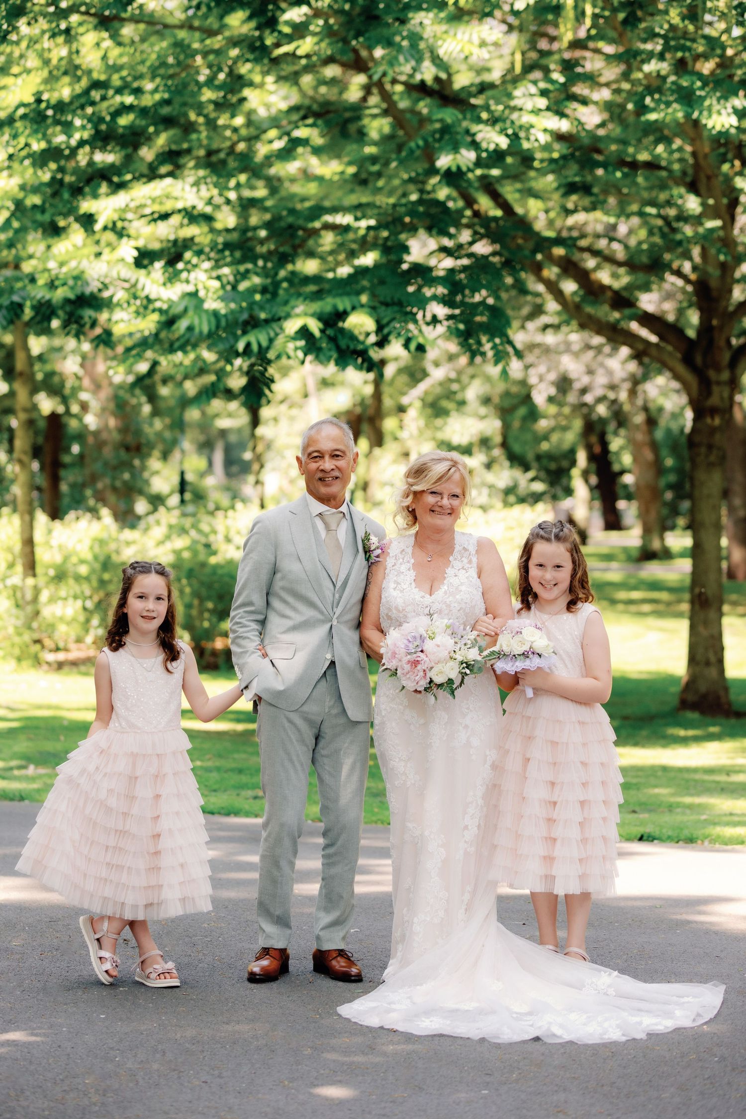 Bride and groom pose with two flower girls in pink dresses in a lush green park setting.