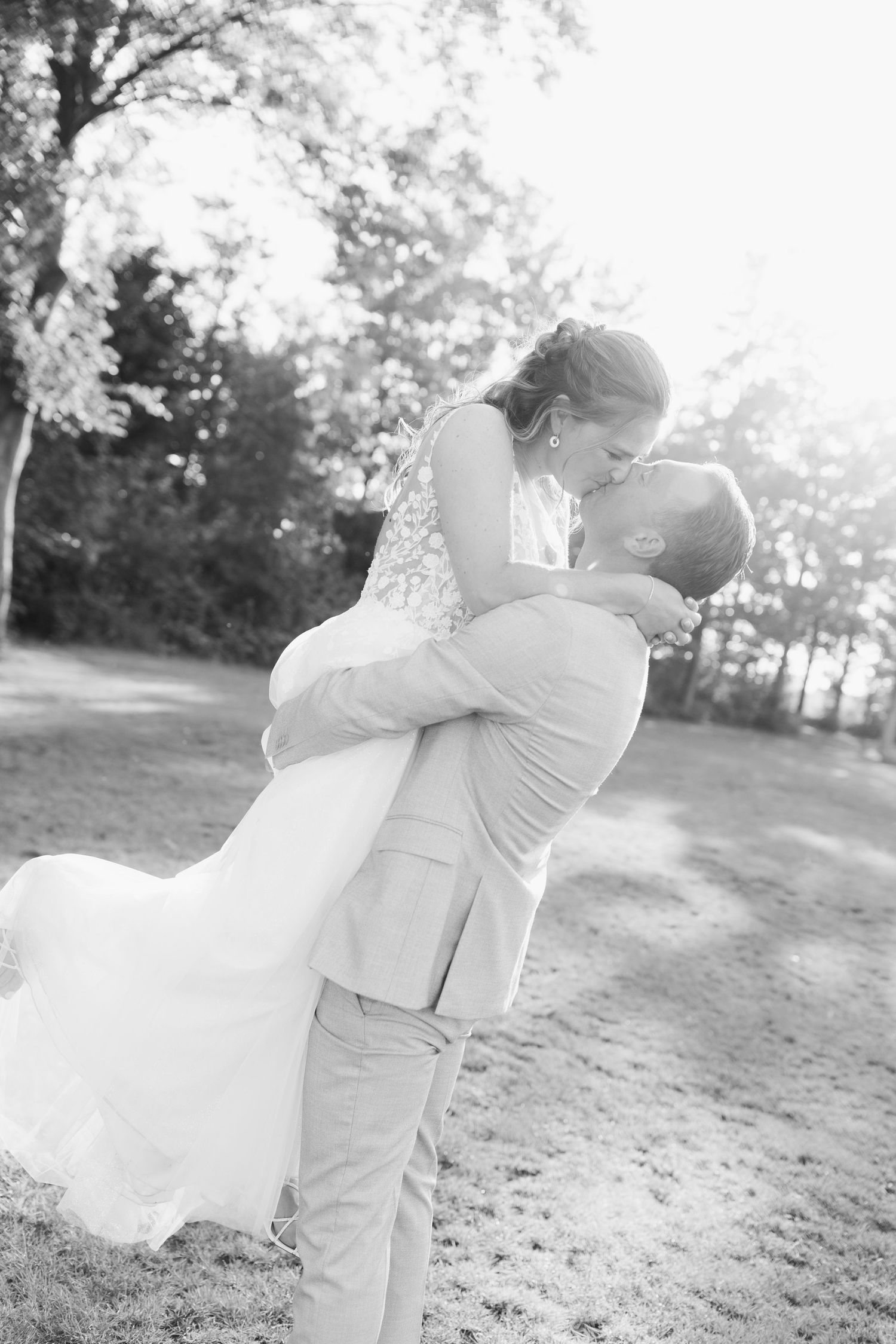 Black and white photo of groom lifting bride for a kiss outdoors, wedding dress flowing, trees in background.