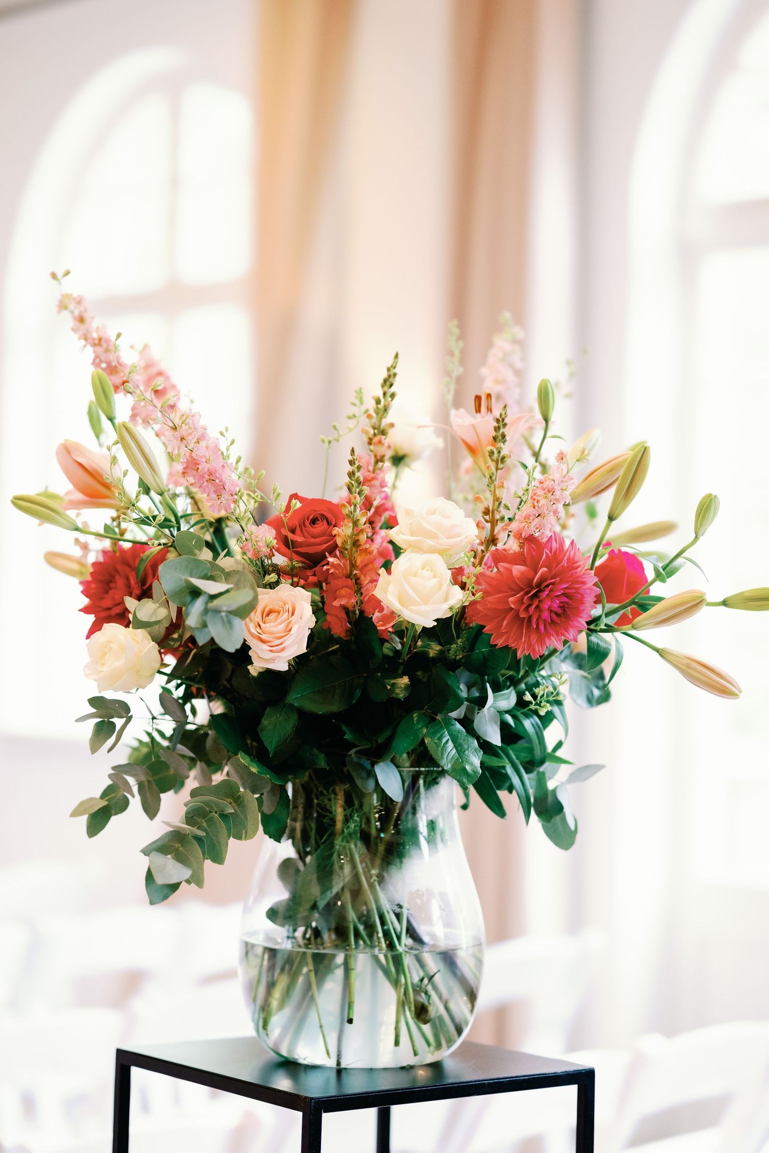 Elegant floral arrangements in glass vases featuring pink and coral blooms with eucalyptus on black stands in a bright room.