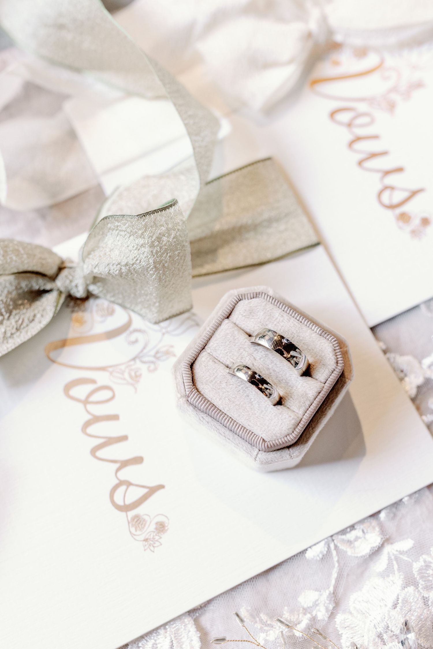 Two silver wedding rings rest in a light gray velvet ring box on a white invitation with elegant script lettering.