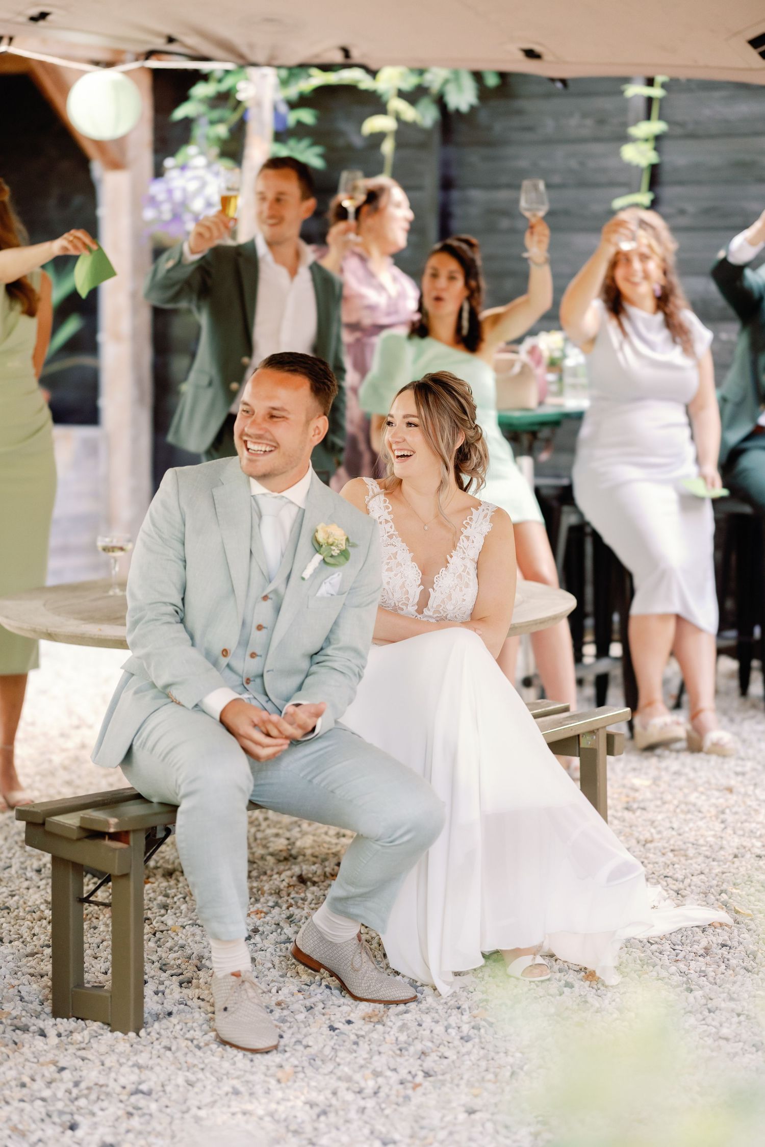 A couple shares joyful laughter while sitting on rustic wooden benches at an outdoor wedding reception.