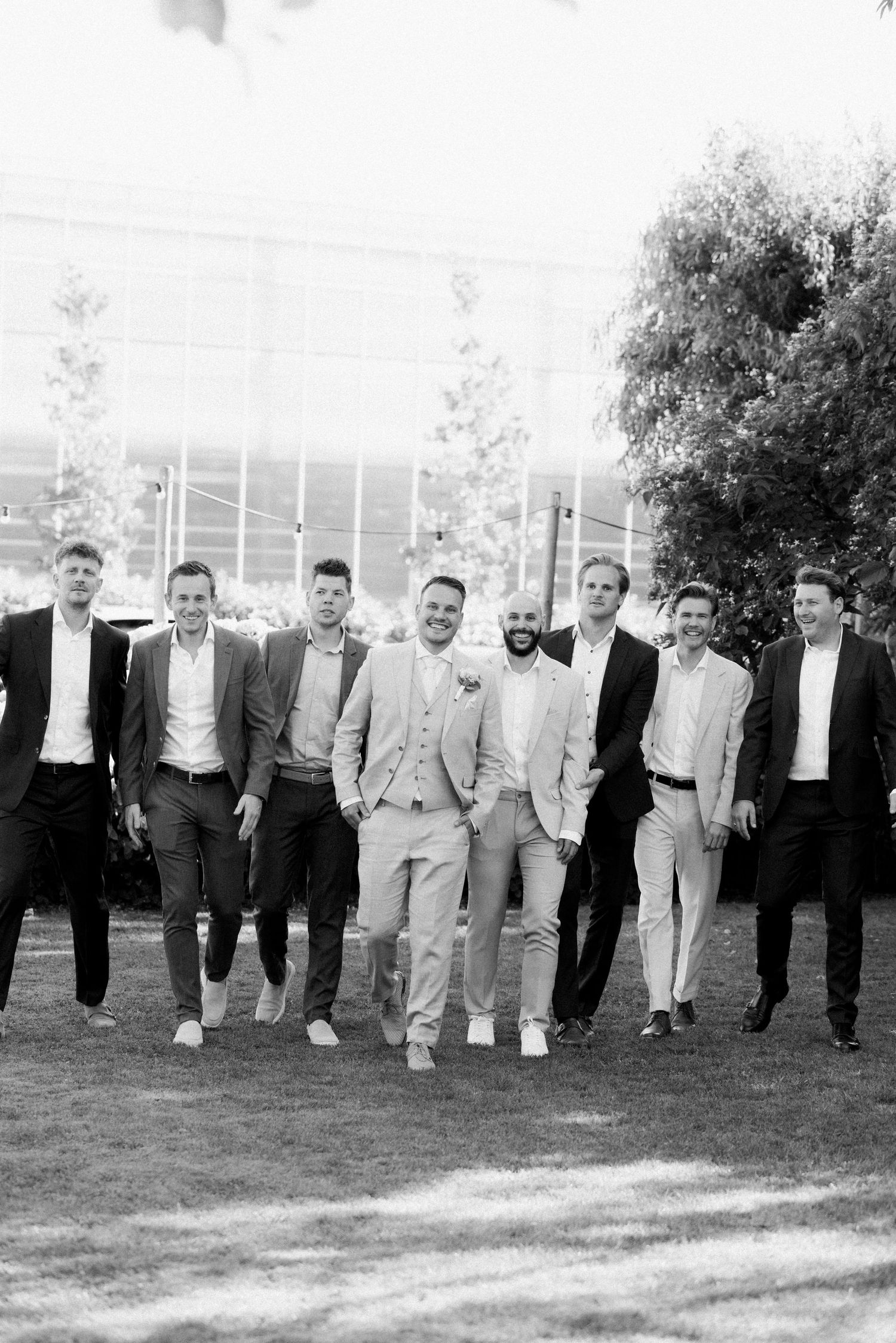 A black and white photo shows groomsmen in suits walking together on a grassy lawn.