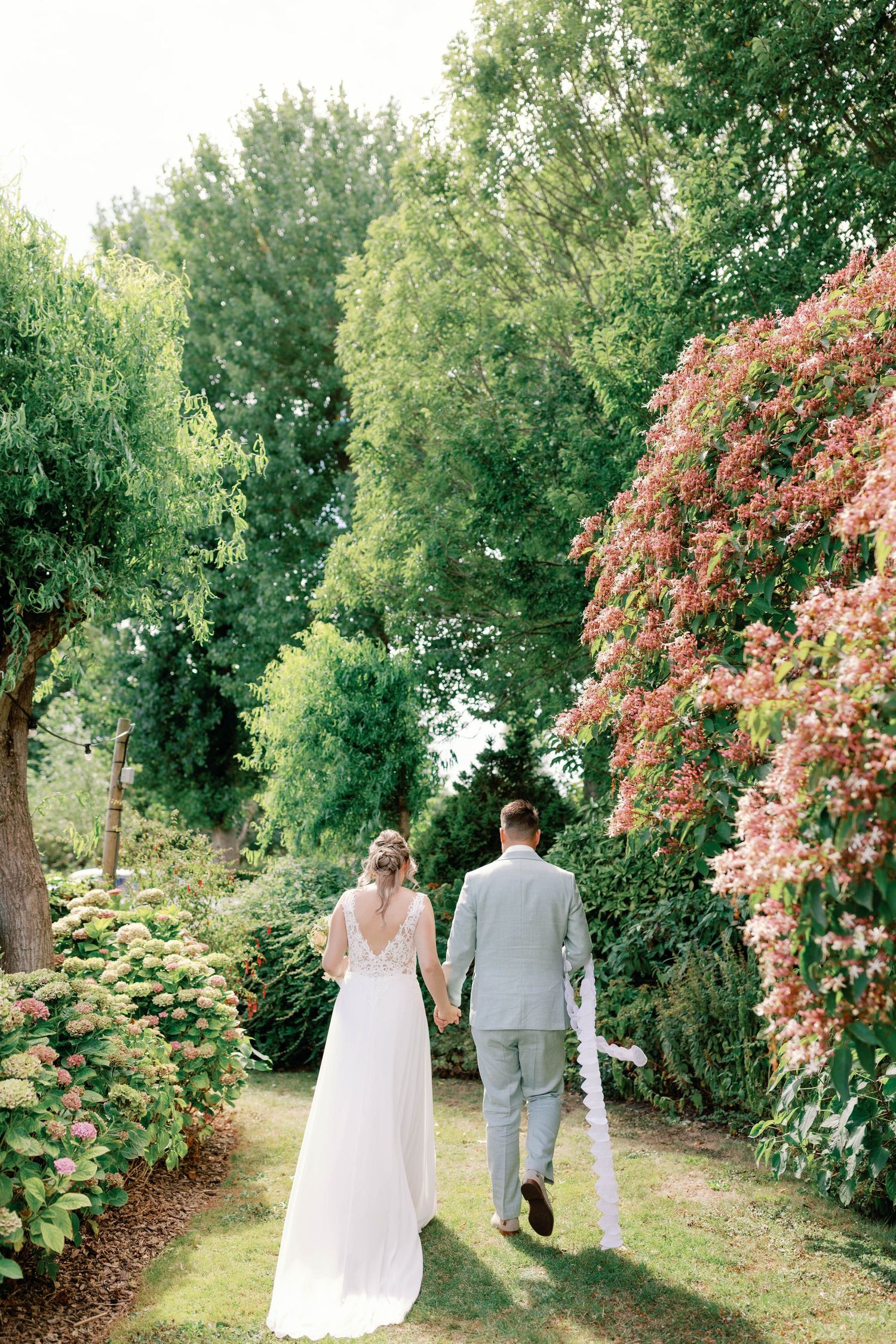 A couple walks hand in hand down a garden path surrounded by lush greenery and blooming pink flowers.