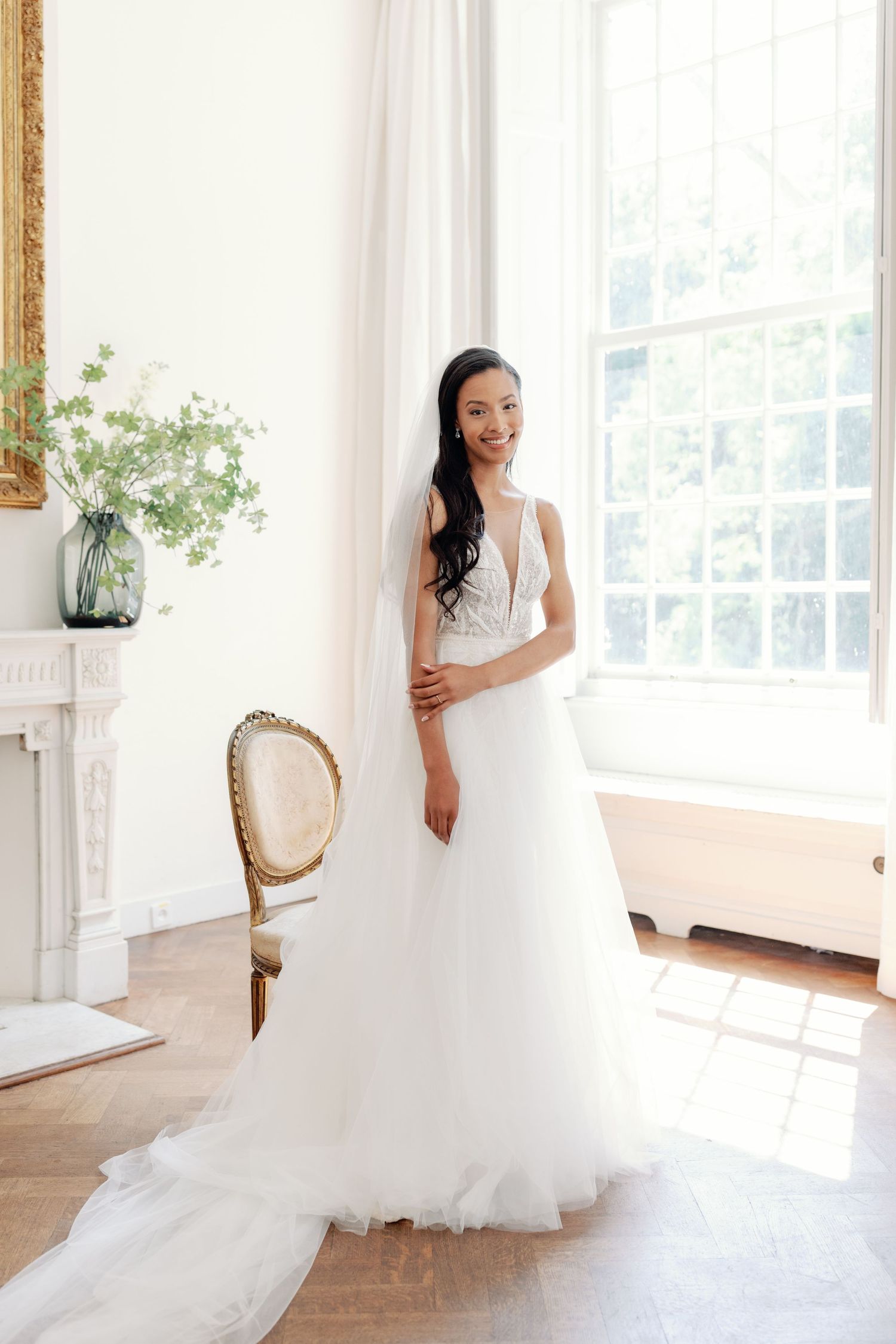 A bride in a flowing white wedding gown twirls gracefully in a bright sunlit room with sheer curtains and hardwood floors.