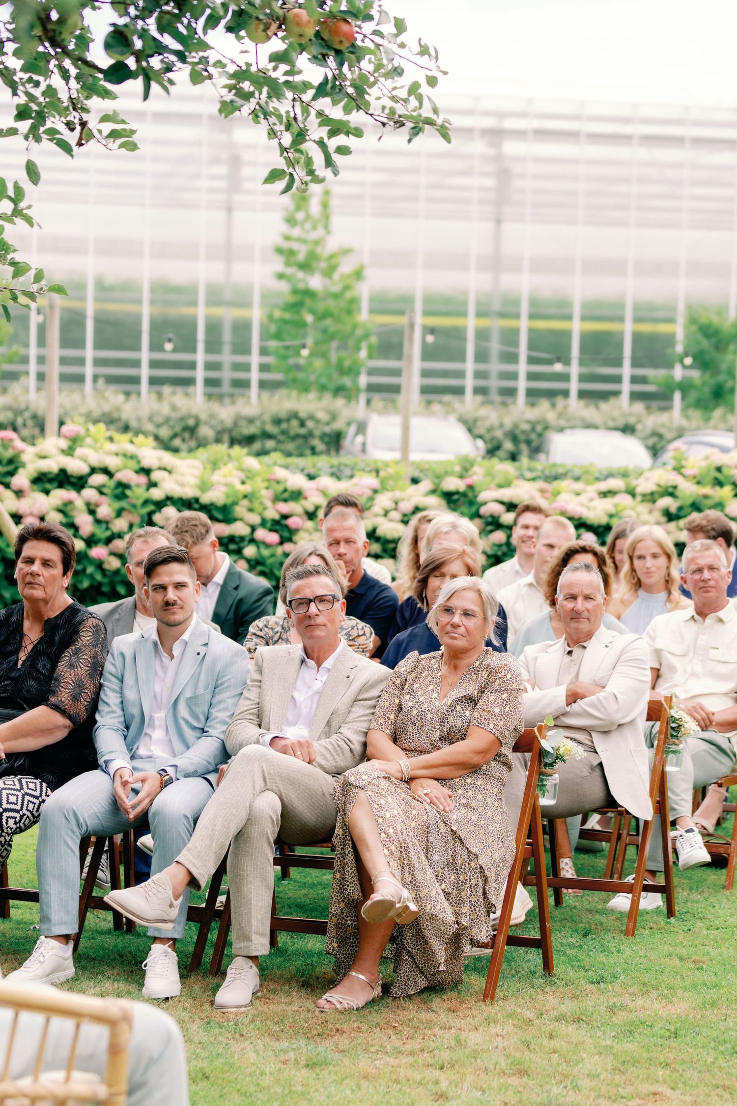 Wedding guests seated on wooden folding chairs on a lawn with a greenhouse in the background during an outdoor ceremony.