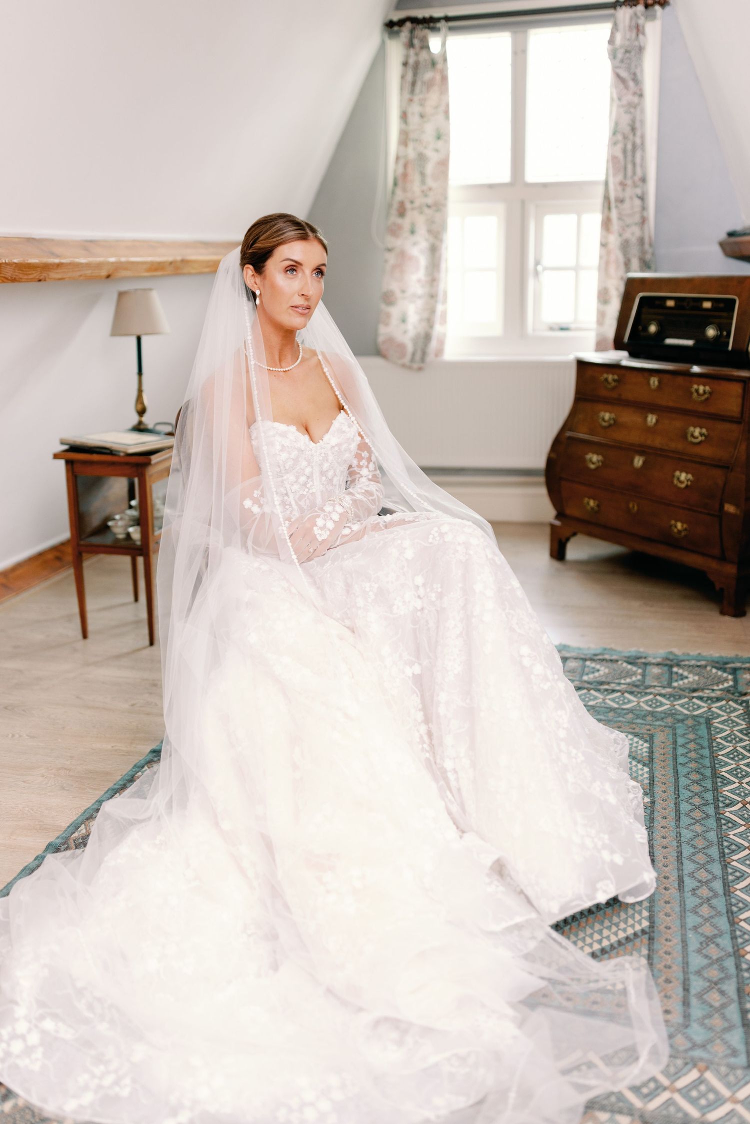 A bride in a flowing white lace wedding gown sits gracefully on a blue patterned rug in a sunlit room.
