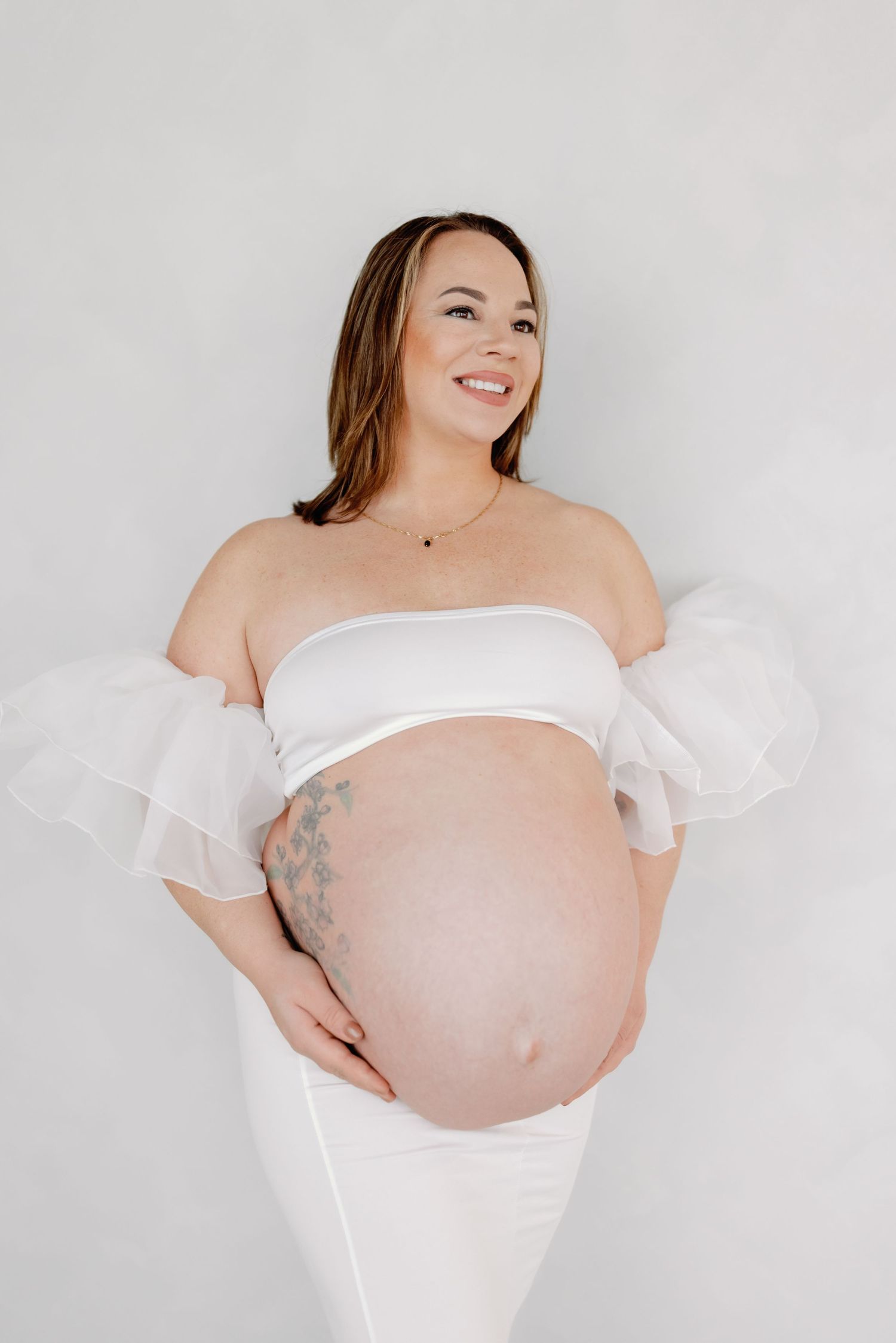Pregnant woman in white off-shoulder dress poses against light background in maternity photoshoot series.