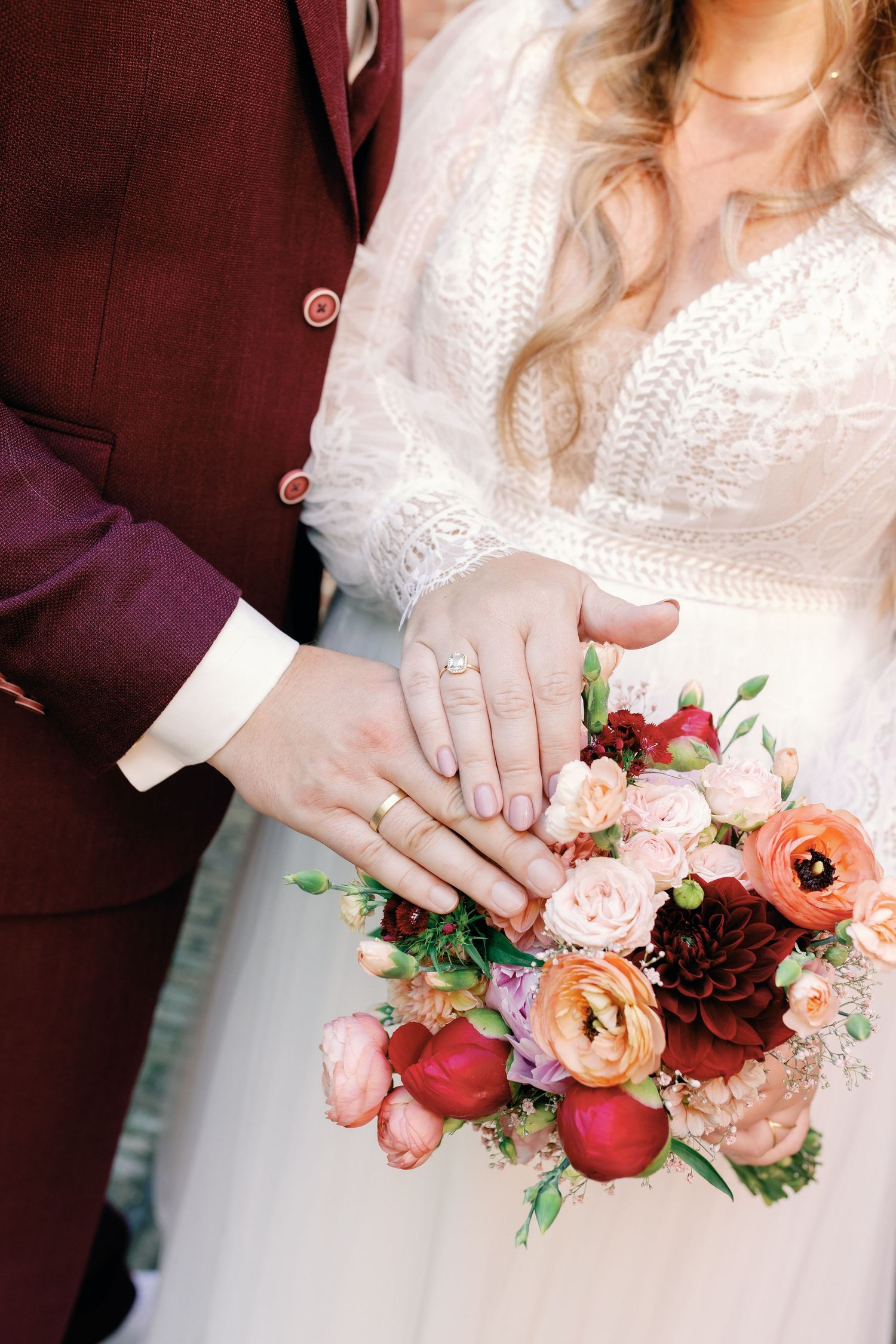 A wedding bouquet featuring peach and burgundy flowers with pine cones is held against a white dress.