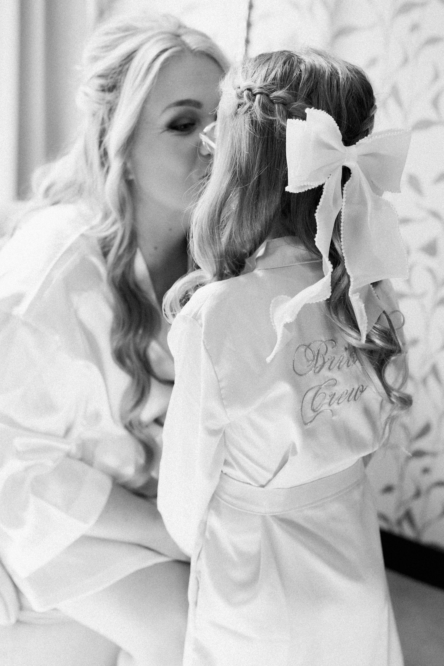 Two women in white robes share a tender moment during wedding preparations.