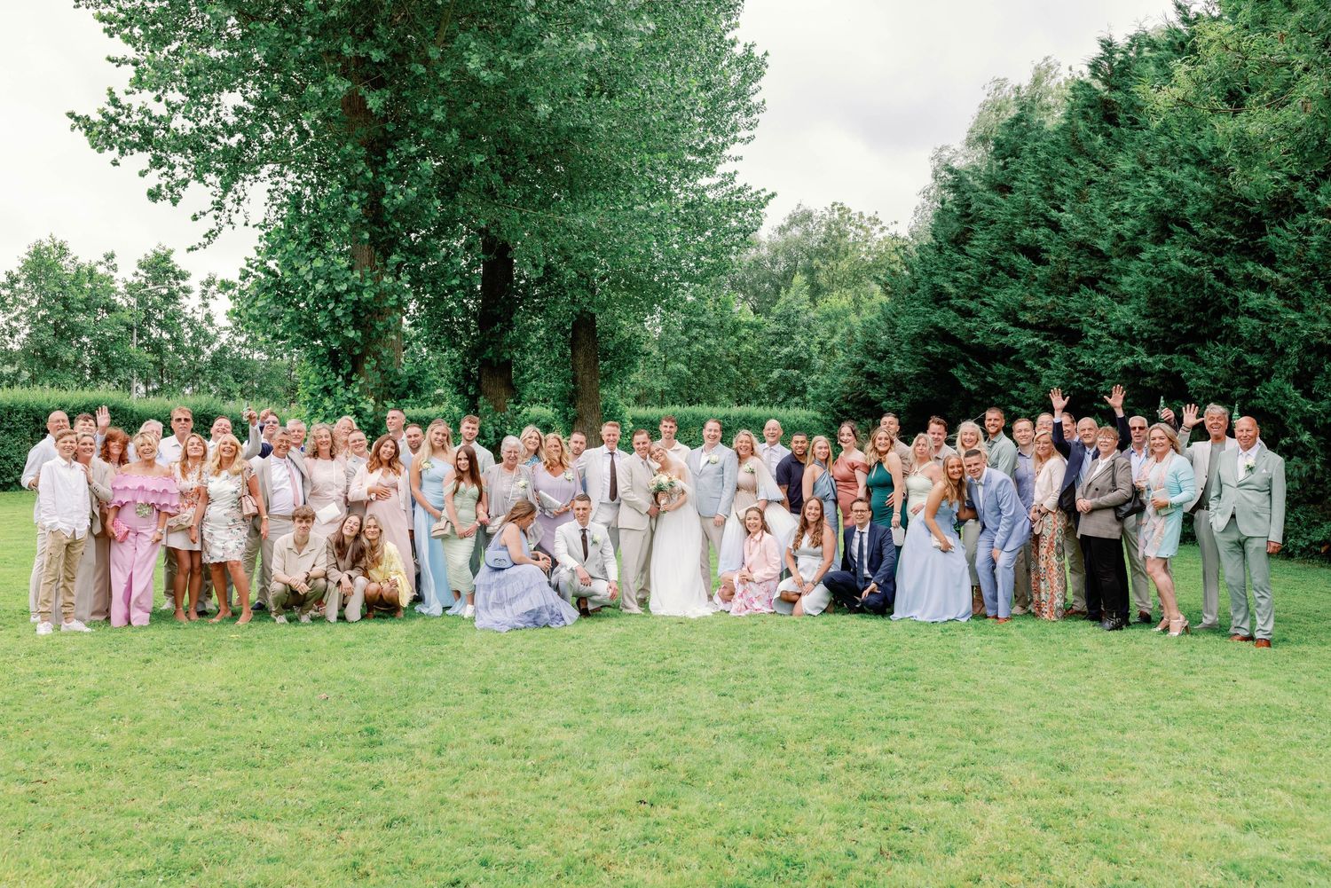 A large wedding party poses together on a grassy lawn surrounded by lush green trees in the background.