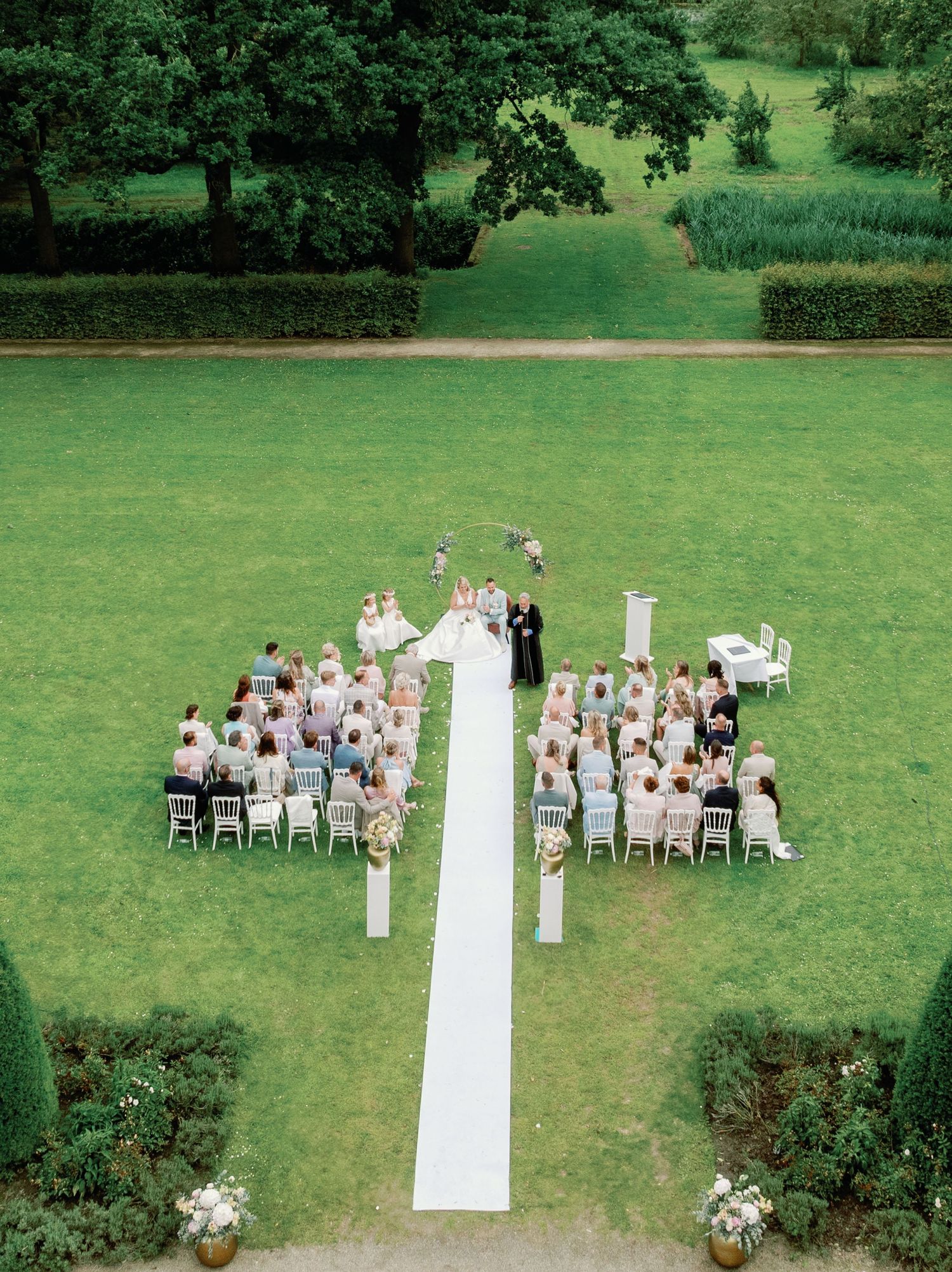 Aerial view of an outdoor wedding ceremony on a manicured lawn with white chairs and floral arrangements along a white aisle runner.
