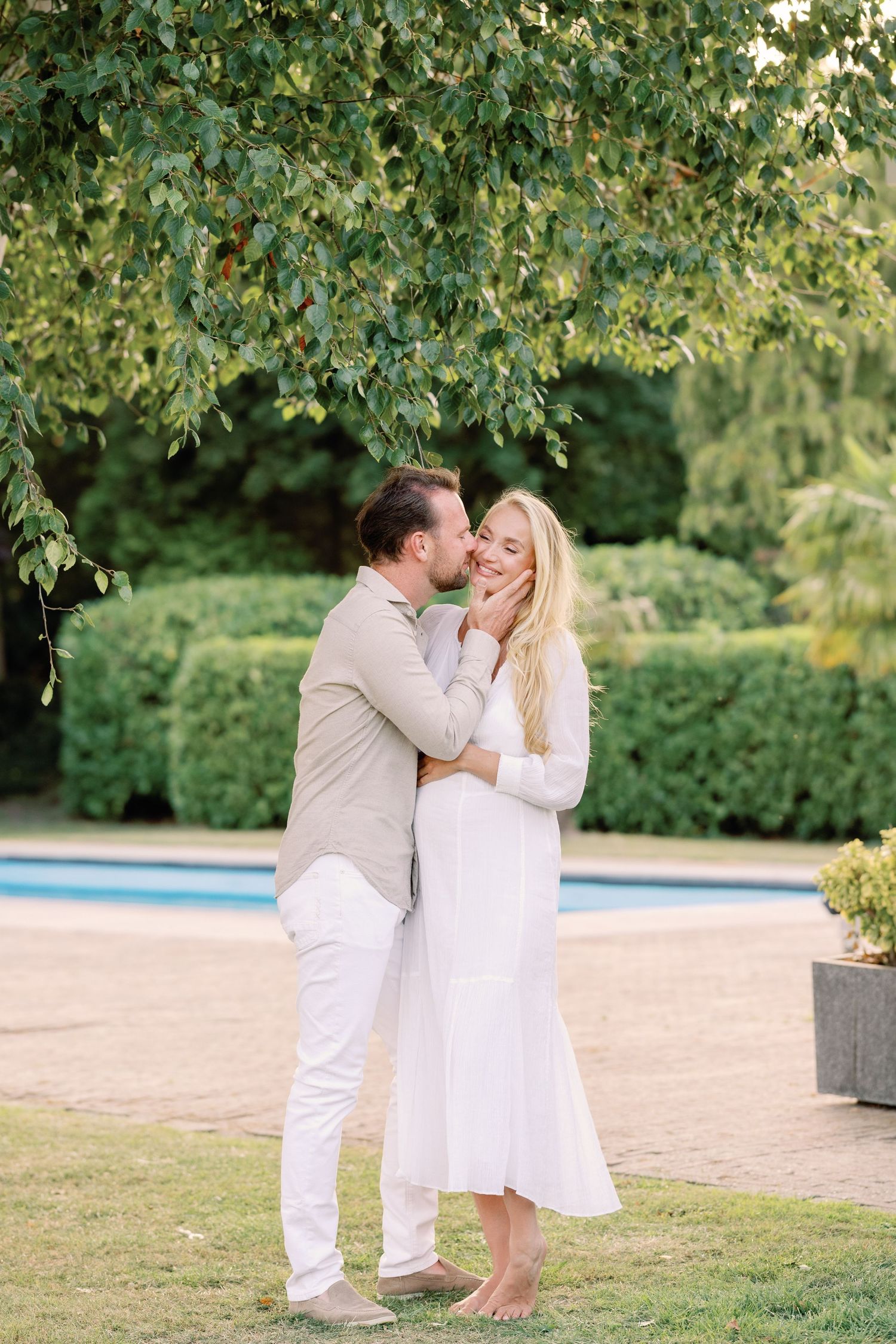 Couple sharing a romantic moment by a pool in a lush garden, man in beige kissing woman in white dress.