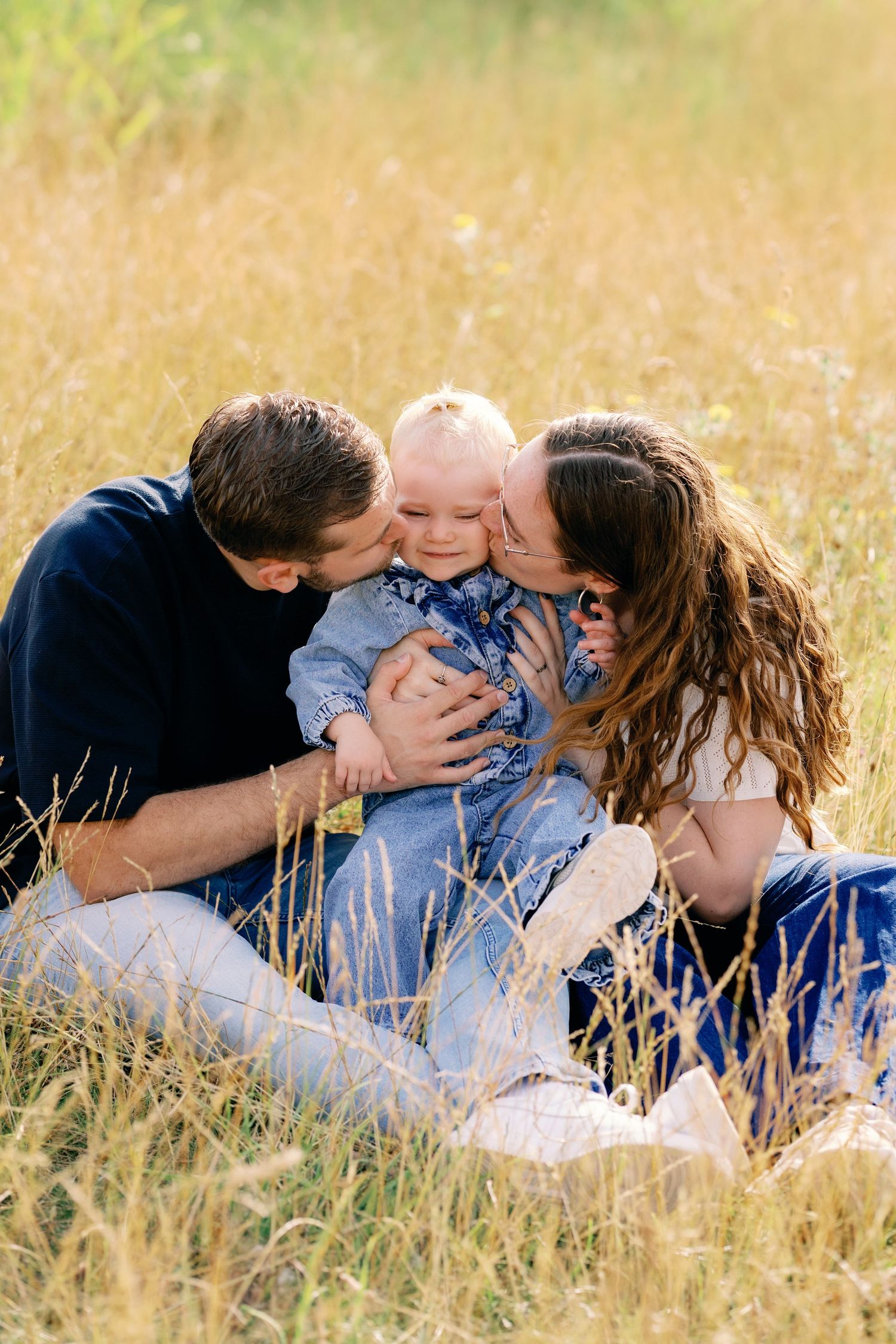 Parents kissing their smiling baby on the cheeks while sitting in a golden field of tall grass on a sunny day.