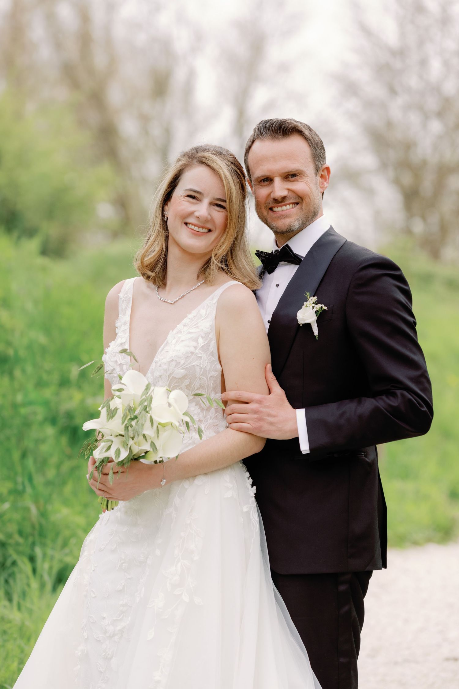 Smiling bride in white lace gown holding bouquet poses with groom in black tuxedo outdoors on wedding day.