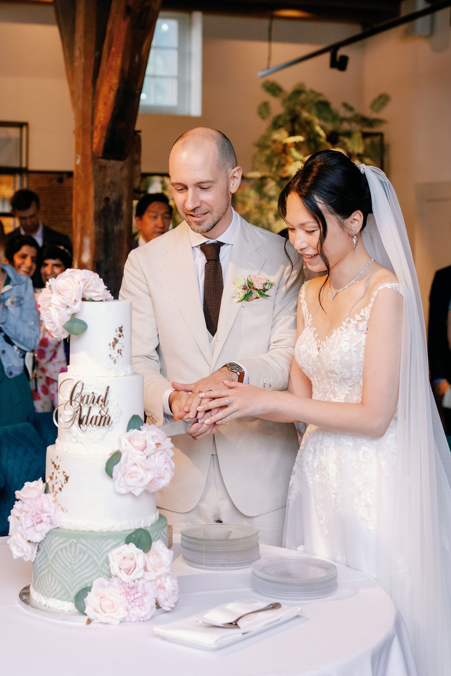 A couple cuts into their elegant white and sage green wedding cake adorned with pink roses at their reception.