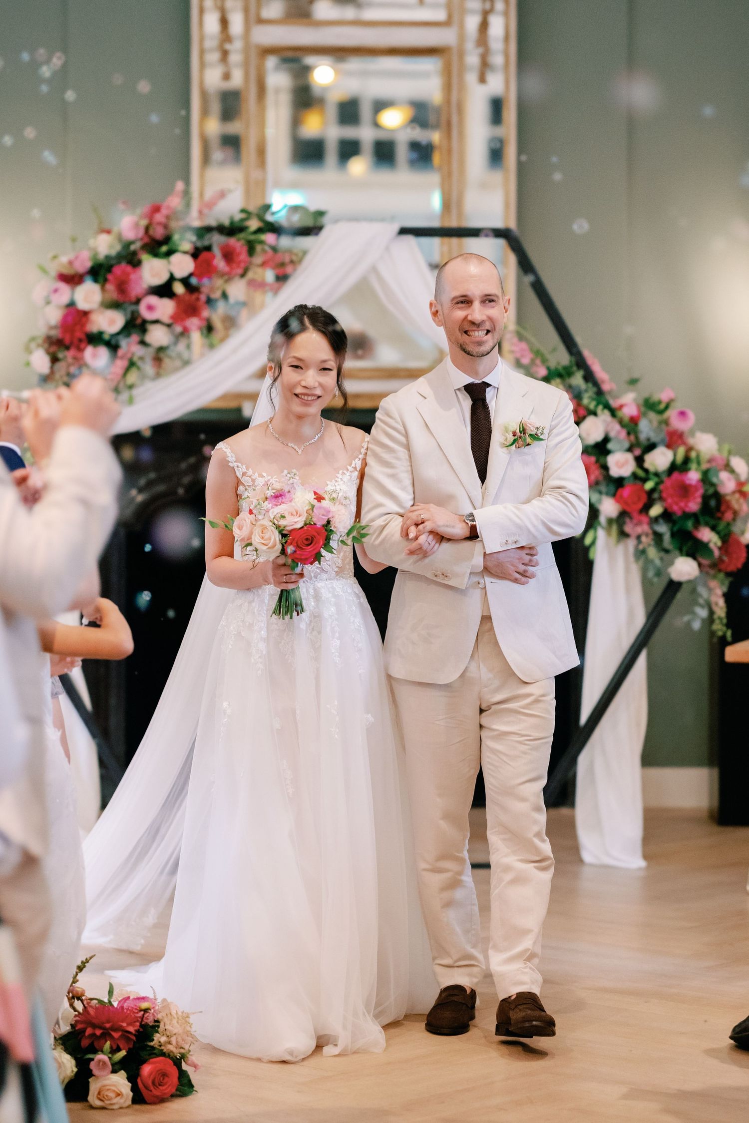 A wedding couple dances together in a bright indoor venue decorated with pink floral arrangements.