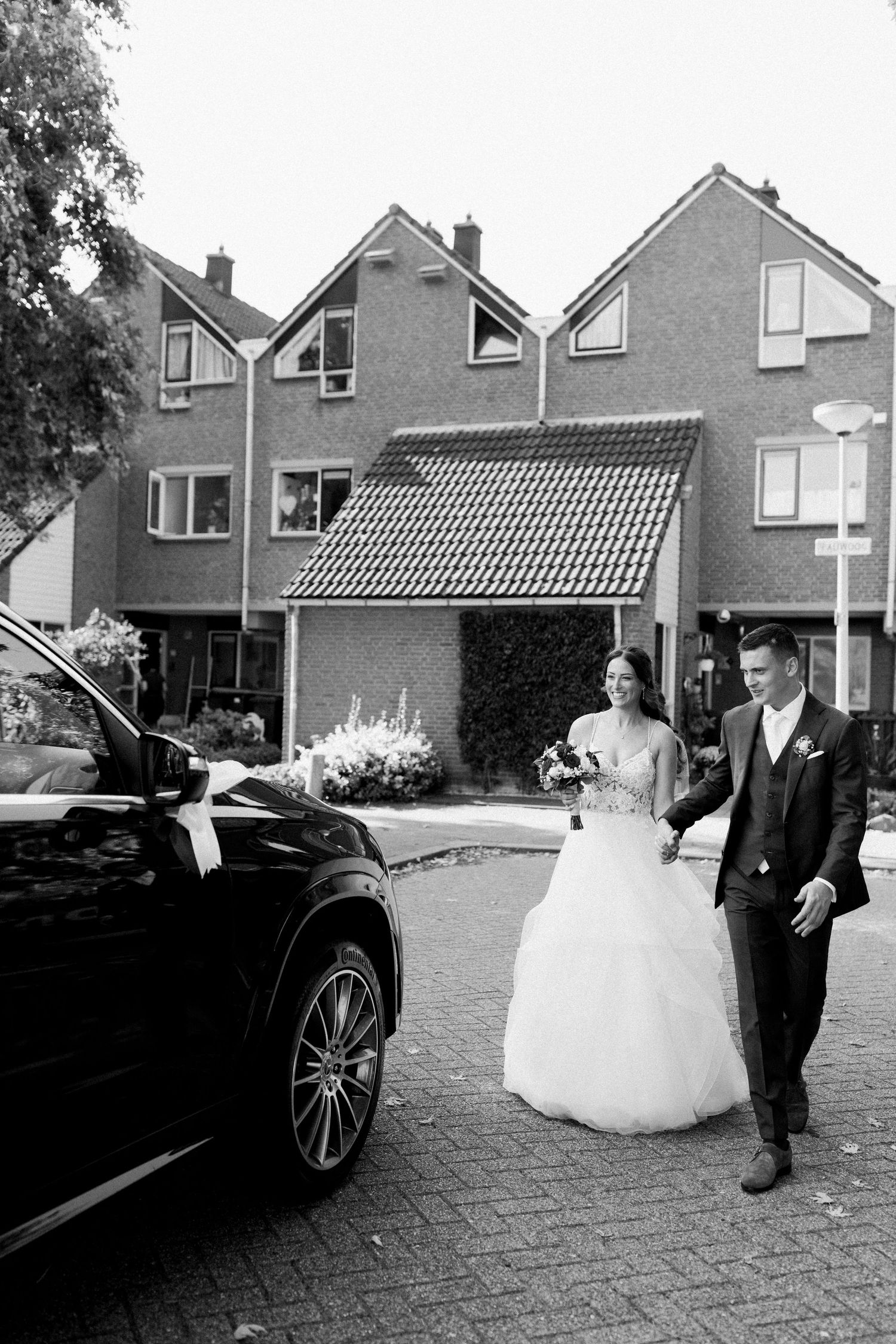 Black and white photo of newlyweds walking near a luxury car outside residential homes.
